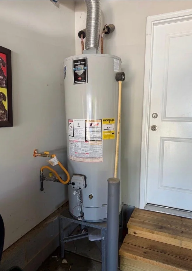 A tall, white water heater with warning labels, located indoors next to a white door and a small wooden platform. It has pipes connected at the top and a yellow flexible pipe on the side.