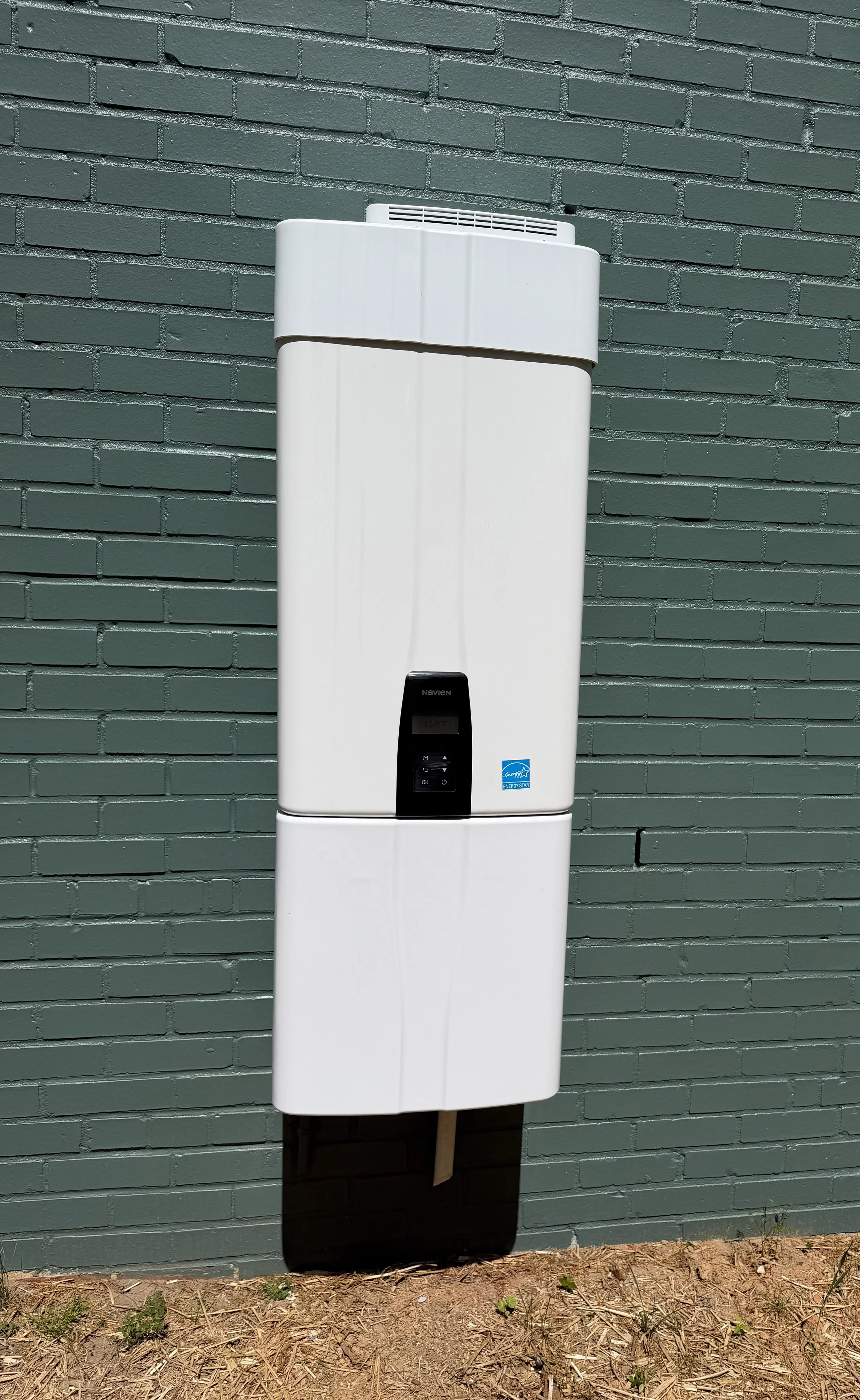 Electric outdoor water heater mounted on a green brick wall outside, with dirt ground and some small plants in front.