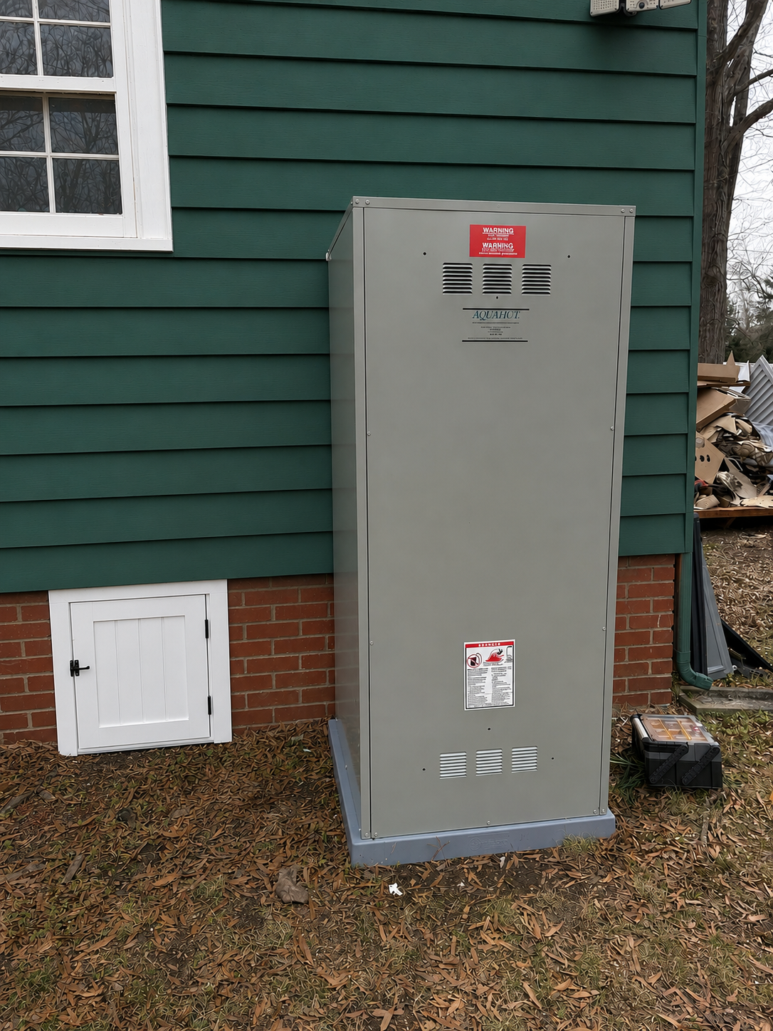Metal electrical equipment box placed outside a house with green siding and a small white door on the wall. The ground is covered with fallen leaves.