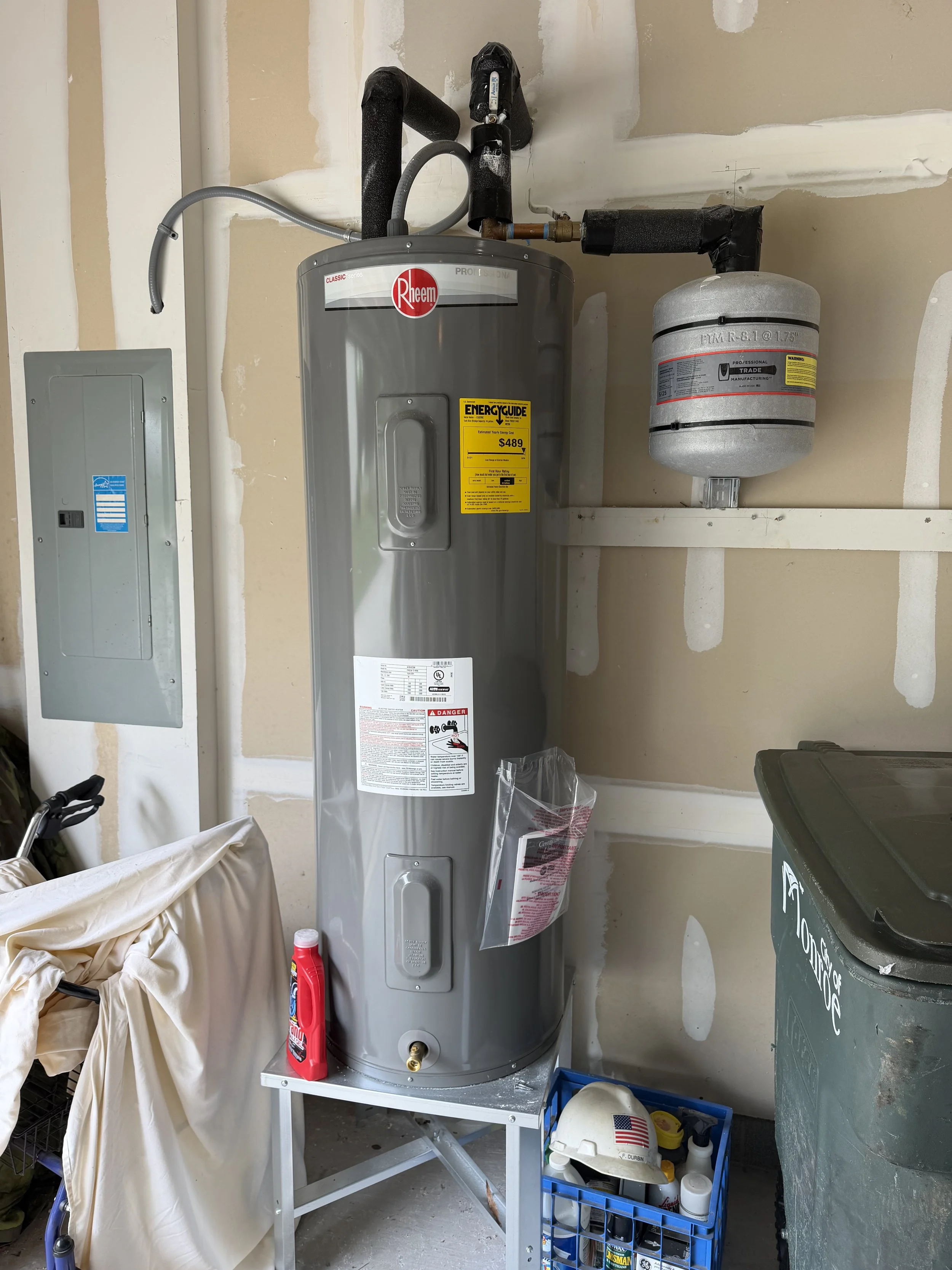 A water heater in a garage next to a trash can, with a helmet and cleaning supplies on a blue plastic crate nearby.
