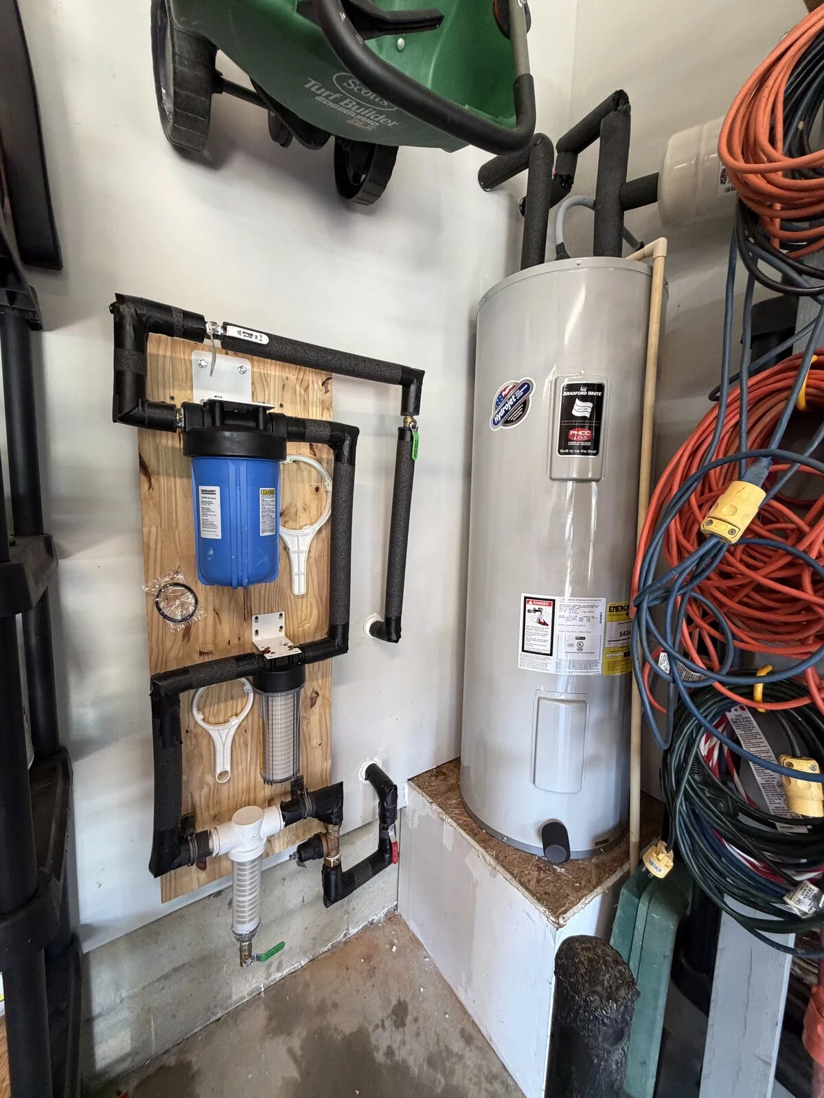 A utility room with a water heater, a mounted water filtration system with blue filter housing, and organized electrical cords on a rack.