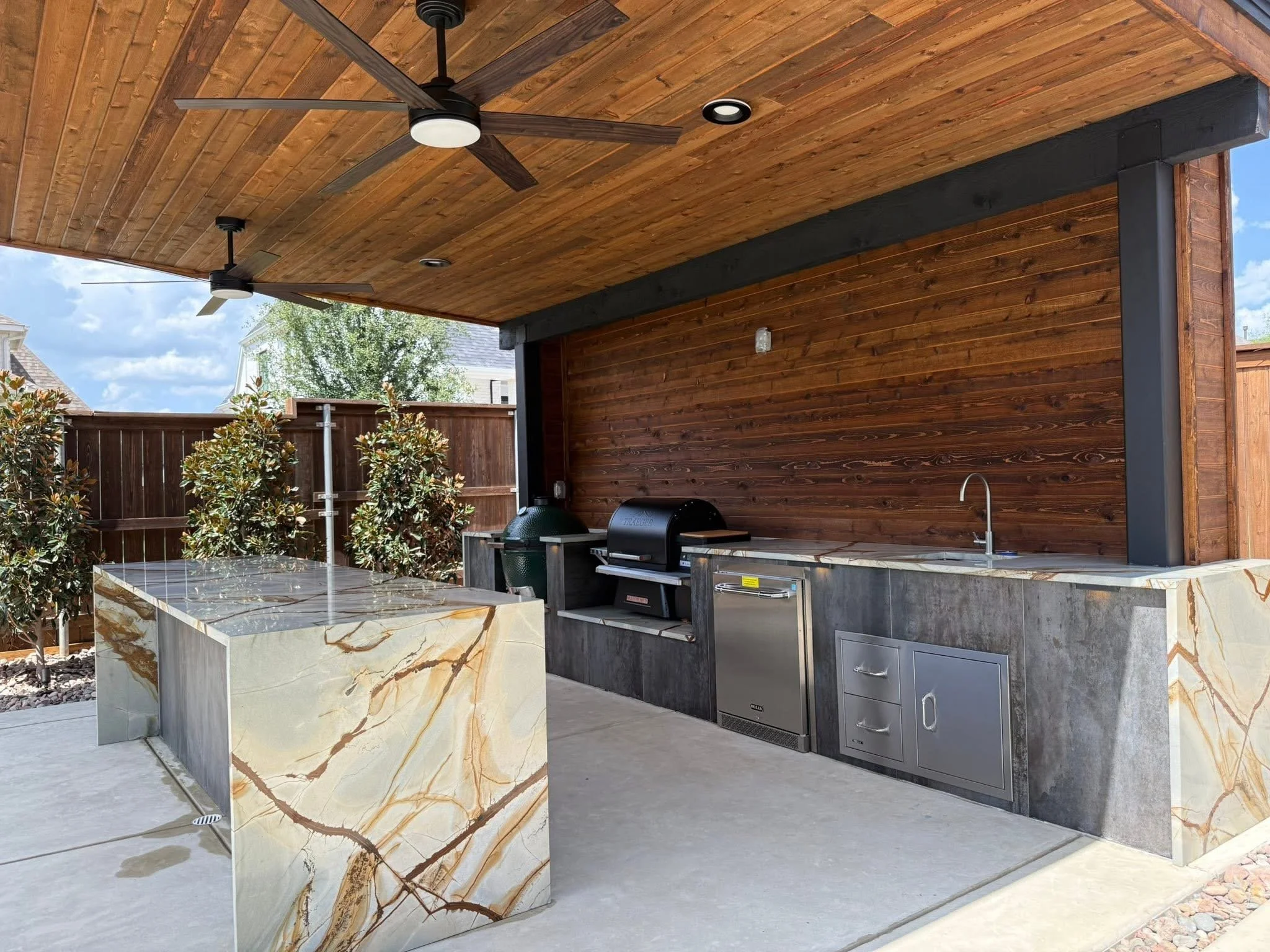 Outdoor kitchen area with a marble countertop, built-in grill, and stainless steel drawers, under a wooden ceiling with ceiling fans and lighting.