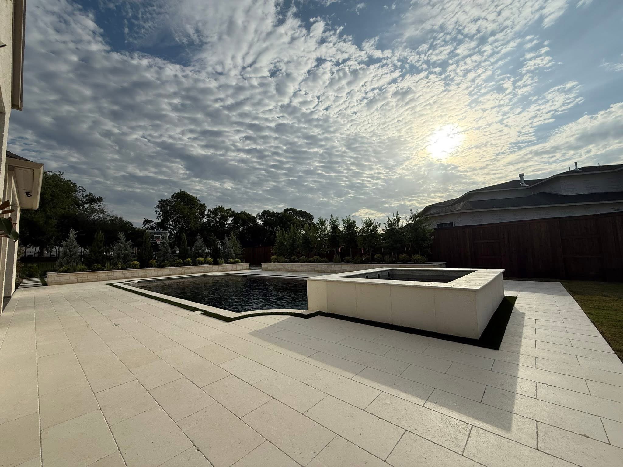 A backyard patio with a water feature, surrounded by trees, with a house on the left and a wooden fence on the right, under a sky with scattered clouds and the sun visible.