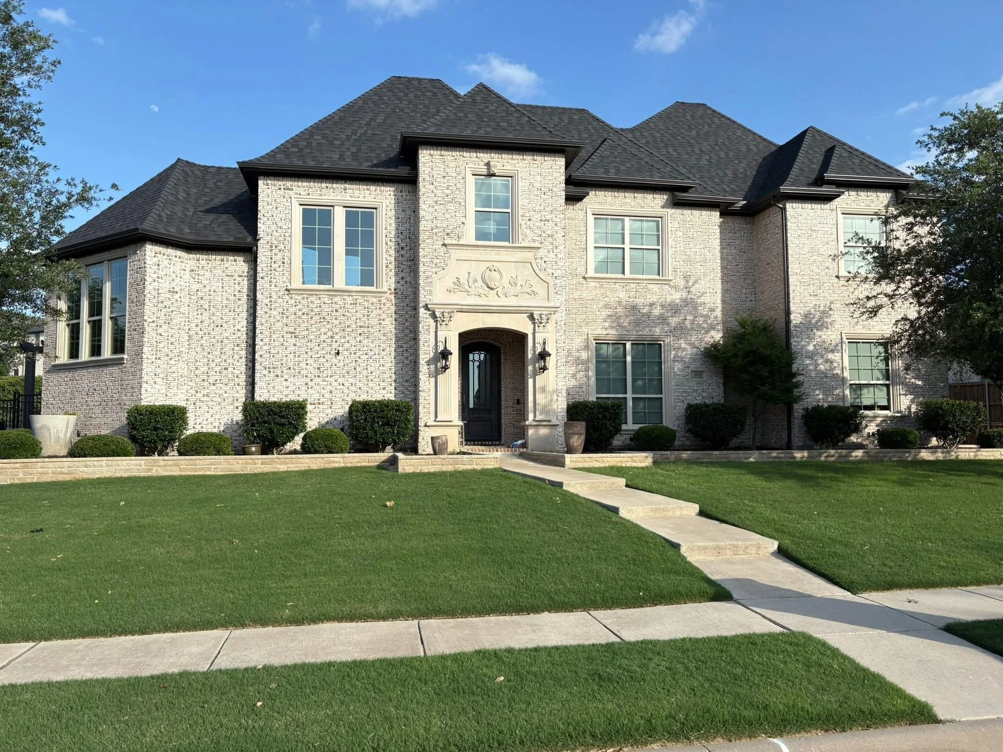 Front view of a two-story house with white brick exterior, black roof, multiple windows, and a small front yard with a sidewalk and lawn.