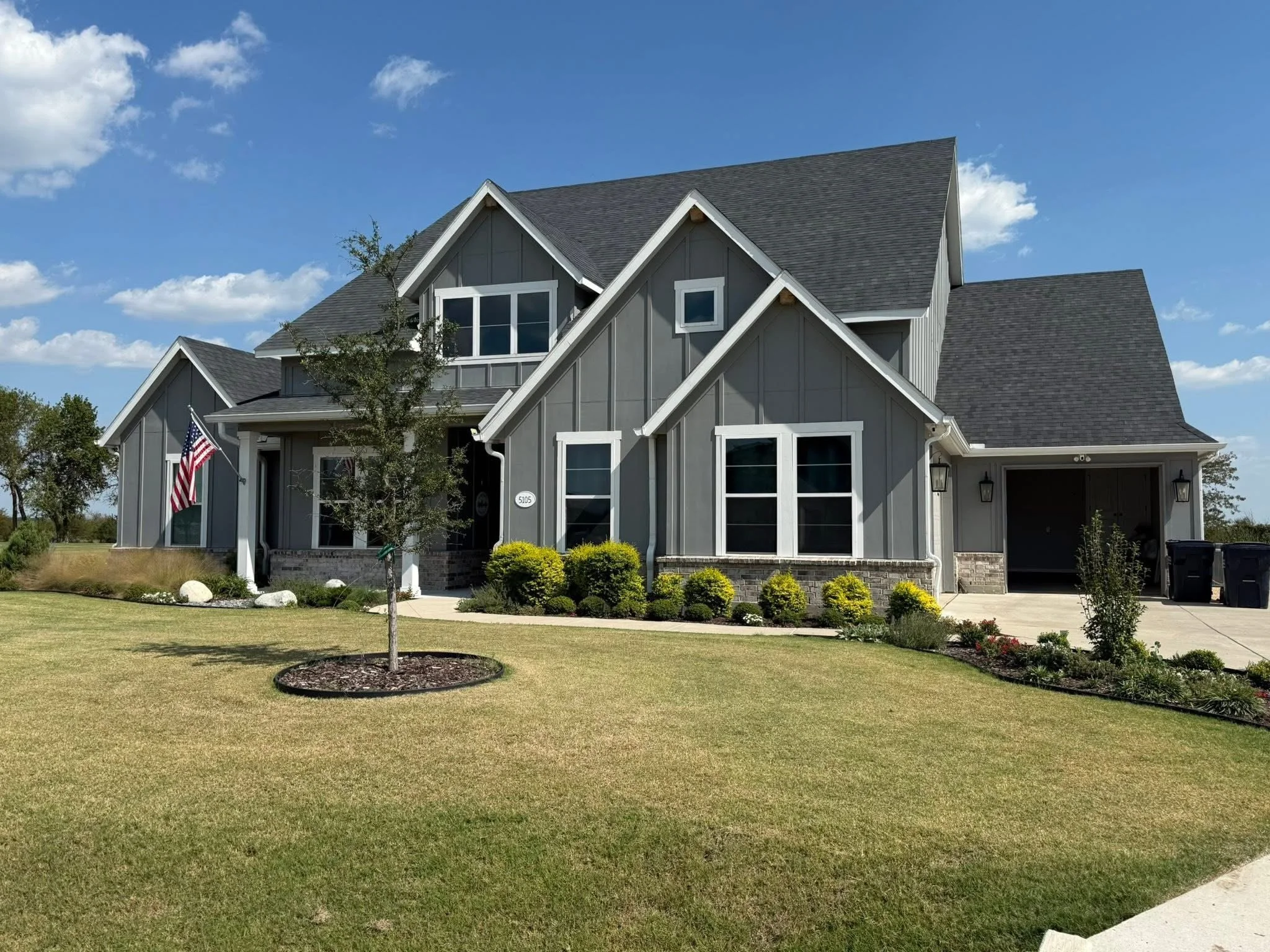 A two-story gray house with white trim, a large front porch, and a sloped roof under a partly cloudy blue sky. The house has multiple gables and large windows. The front yard features a lawn with small bushes, a young tree, and a sidewalk.