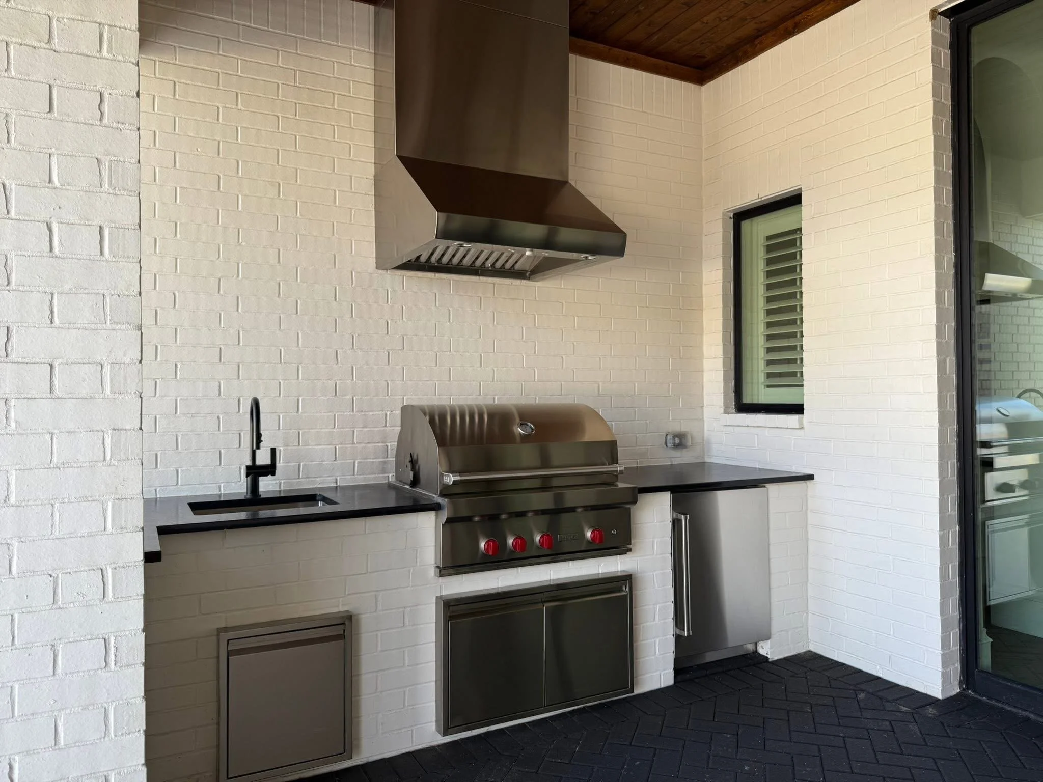 A freshly cleaned  outdoor kitchen with a stainless steel grill, black countertop, white brick wall, and large windows.