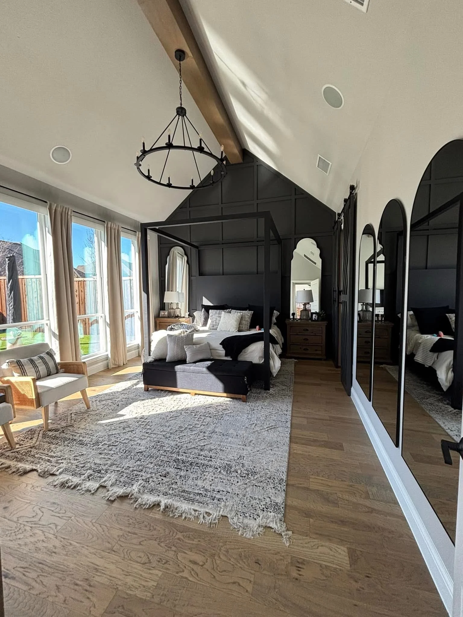 A bedroom with a black canopy bed, wooden nightstand with lamp, and three mirrors. Large windows with beige curtains allow sunlight to fill the room. A patterned rug covers part of the wooden floor.