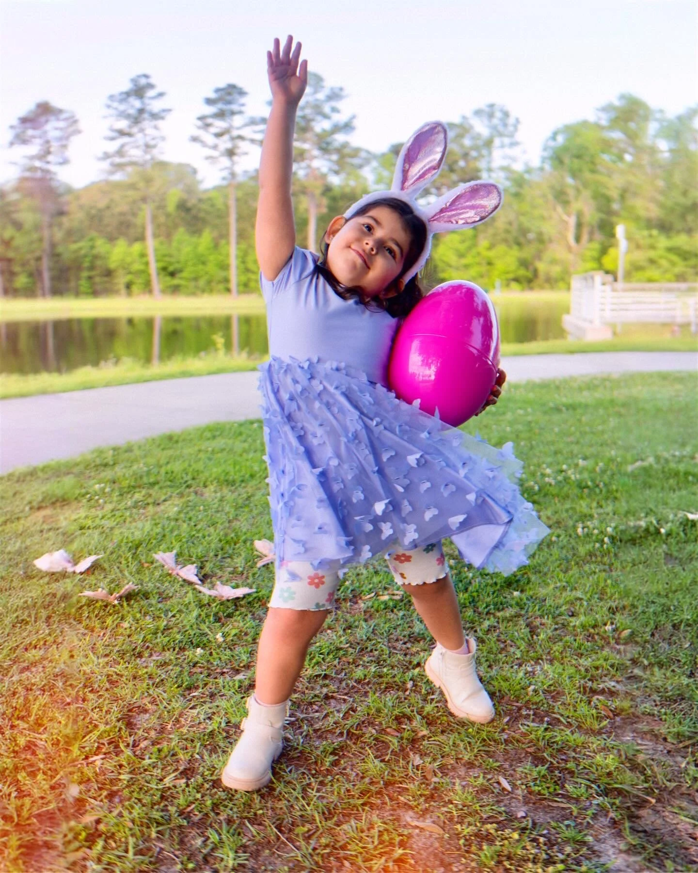 Young girl dressed as a bunny holding a pink Easter egg outdoors near a lake with trees in the background.
