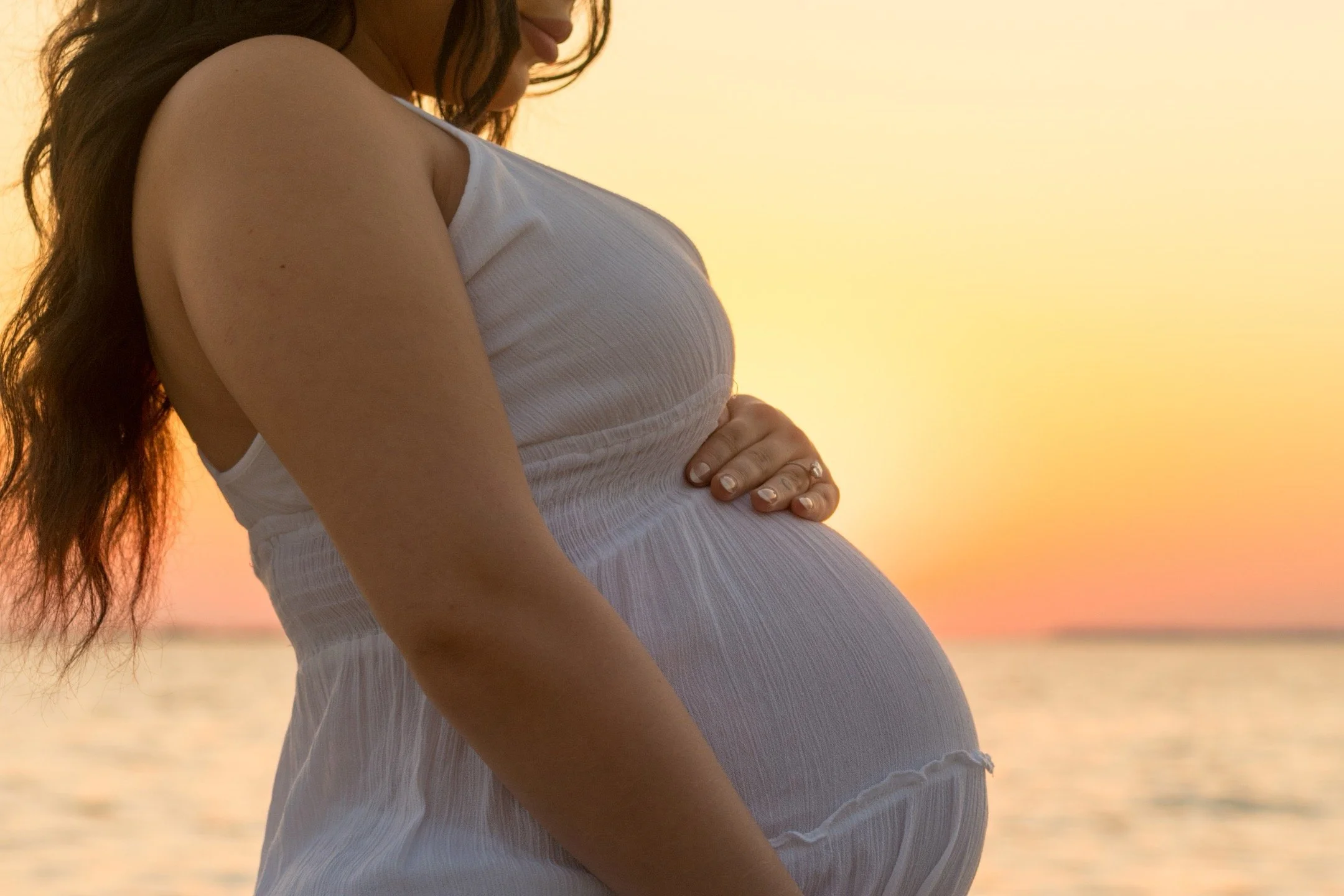 A pregnant woman in a white dress standing by the water during sunset