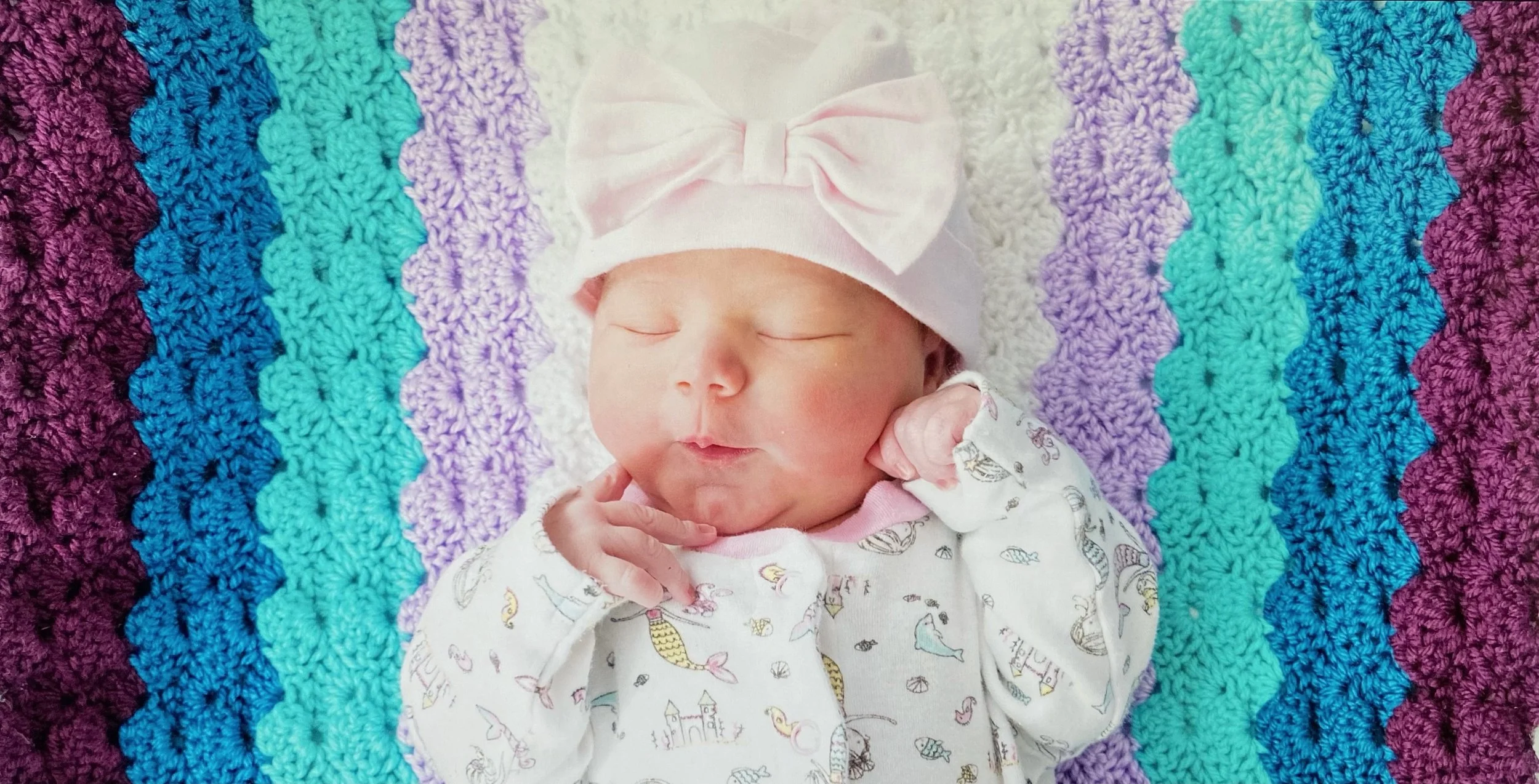 A sleeping baby with a pink bow hat lying on a multicolored crochet blanket with purple, turquoise, and white zigzag patterns. The baby wears a white onesie with a nautical theme.