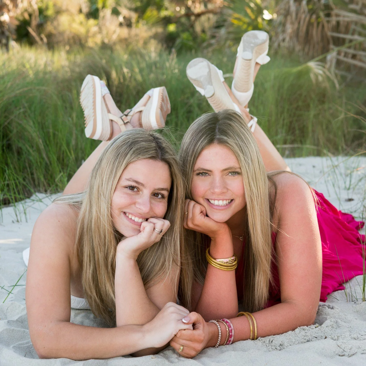 Two young women lying on the sand at the beach, smiling, with grass and trees in the background.