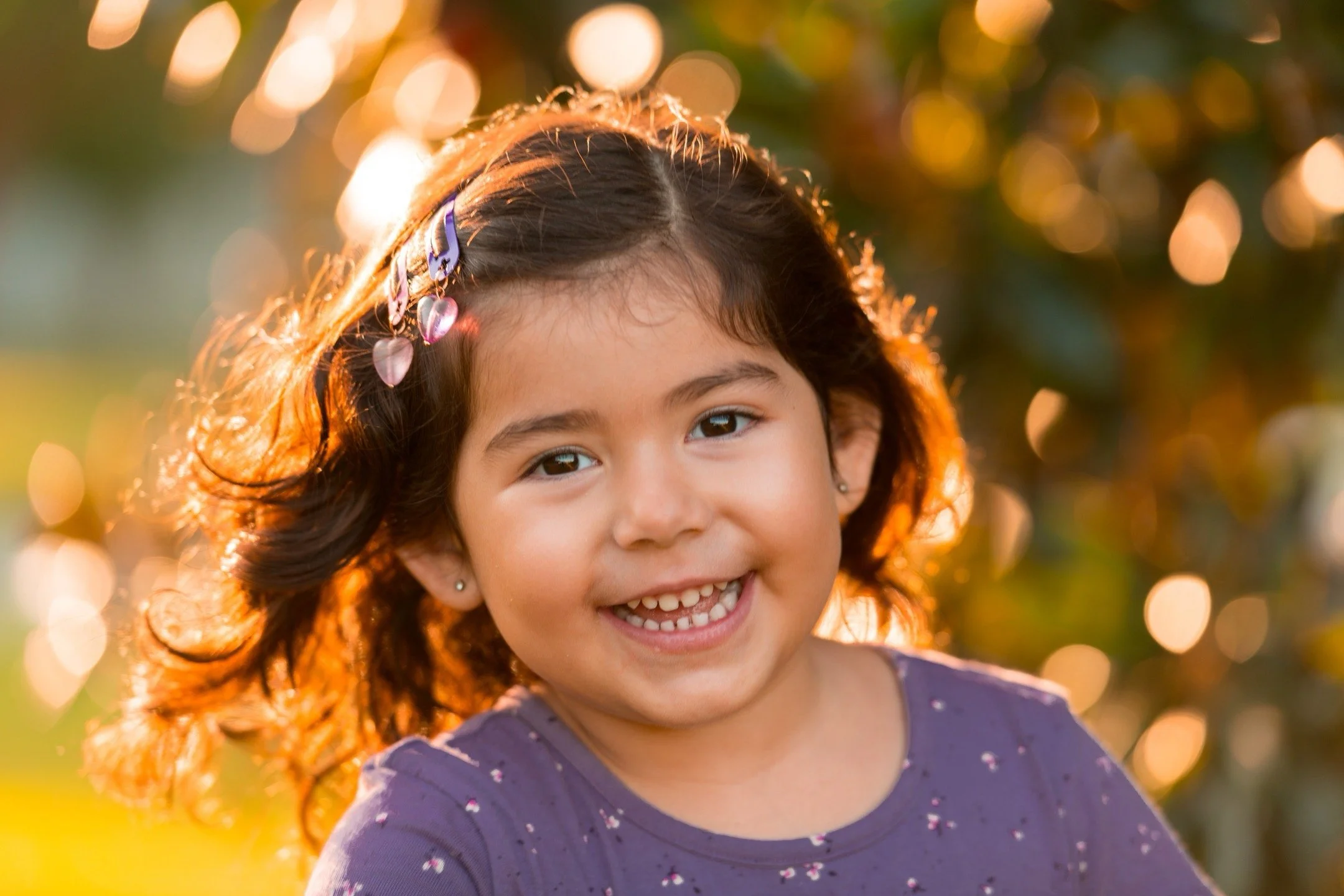 A young girl with dark hair smiling outdoors with sunlight backlighting her, wearing a purple shirt with small details and hair clips with pink hearts.