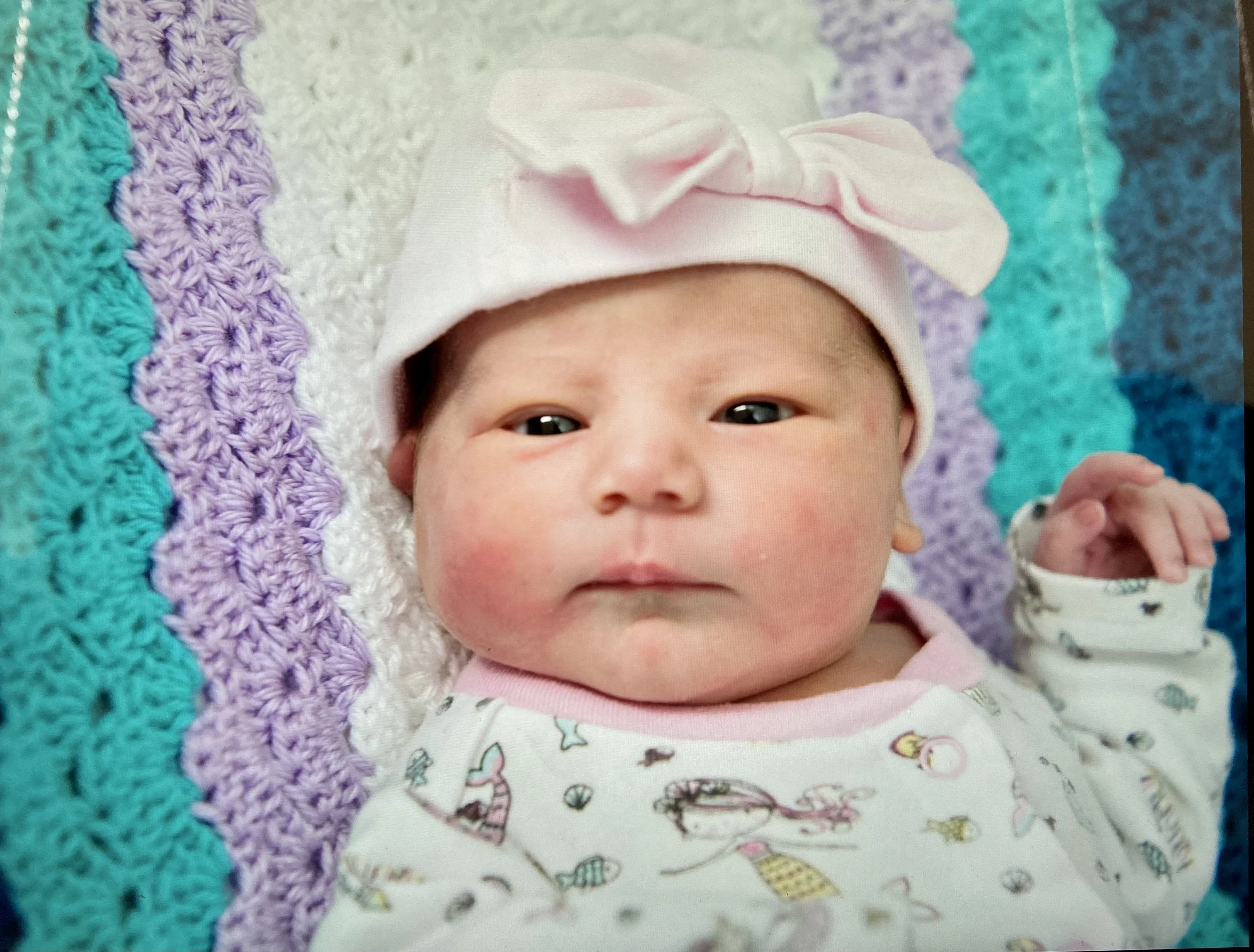 Close-up of a baby wearing a white hat with a pink bow and a white onesie with colorful prints, lying on a multicolored crocheted blanket.