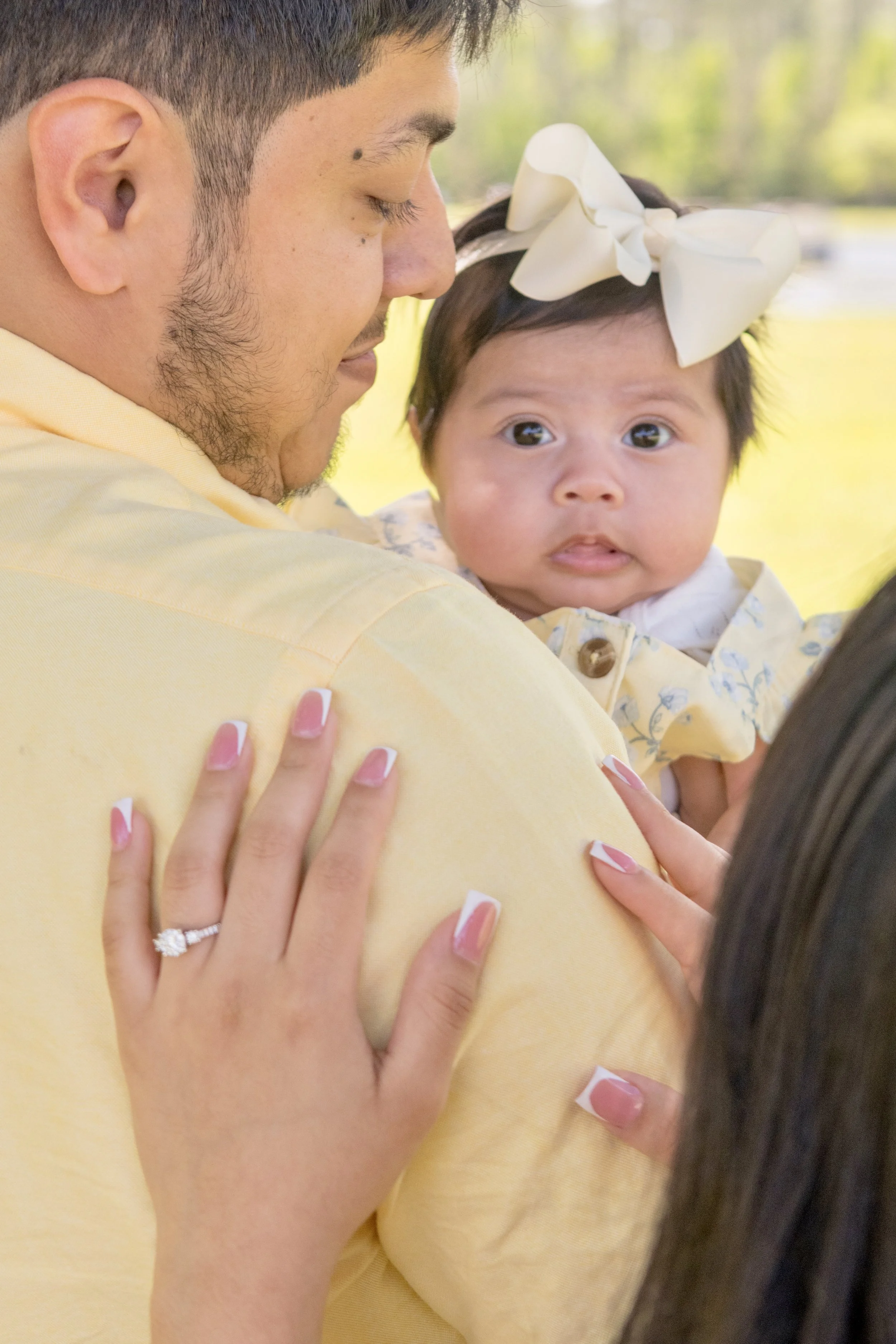 A man holding a baby girl outside in a park, the man has his eyes closed and the baby is looking at the camera with wide eyes, a woman with manicured nails and an engagement ring touches the man's shoulder.