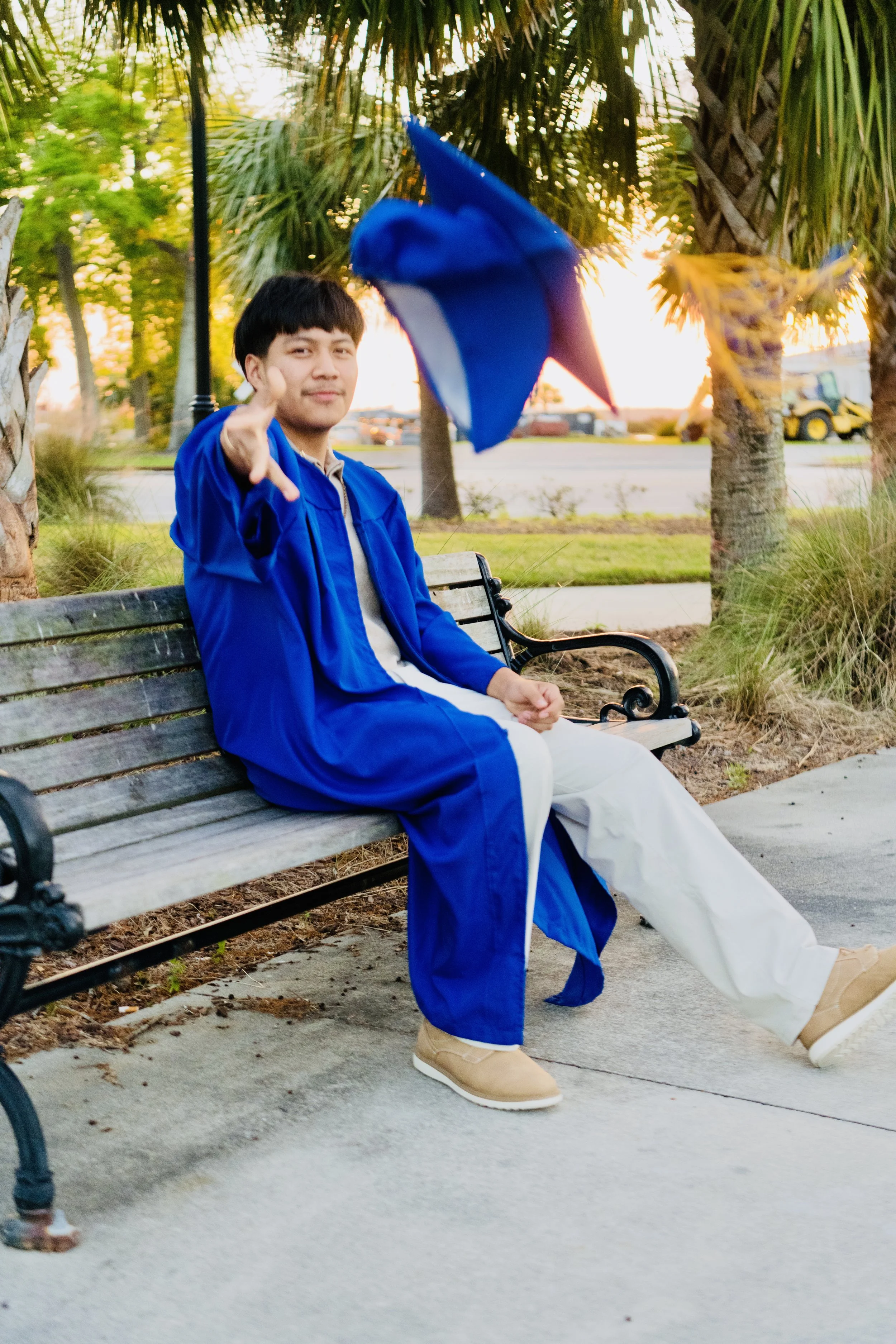 A young man in a blue graduation gown sitting on a park bench, tossing a blue graduation cap into the air during sunset.