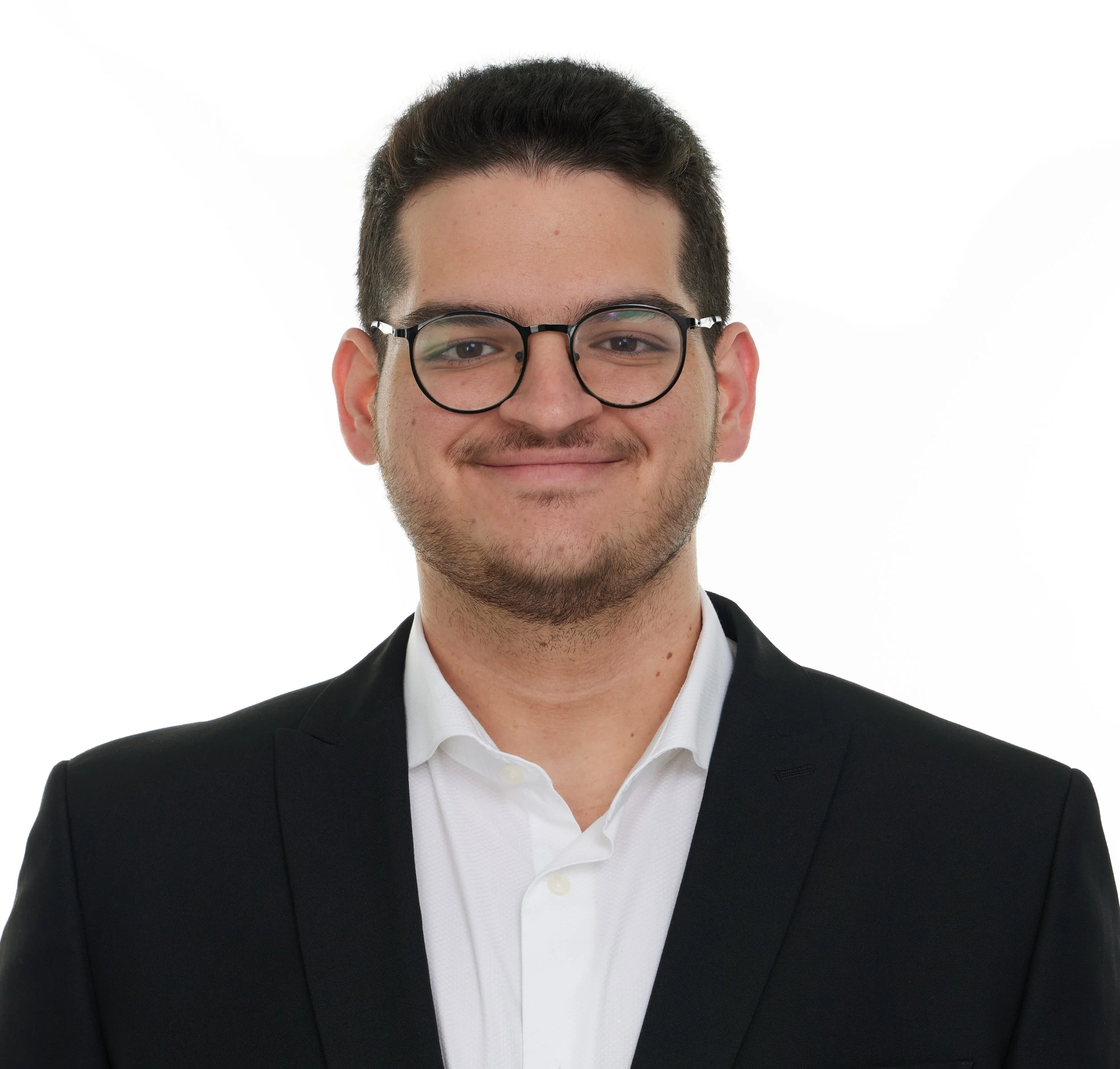 Close-up of a young man with short dark hair, glasses, and light facial hair, smiling and wearing a black suit and white shirt against a white background.