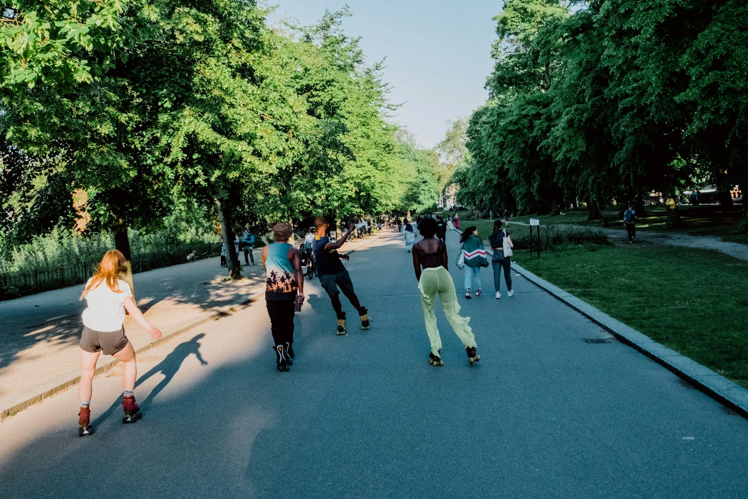 People roller skating and walking in a park on a sunny day, lined with green trees and a paved pathway.