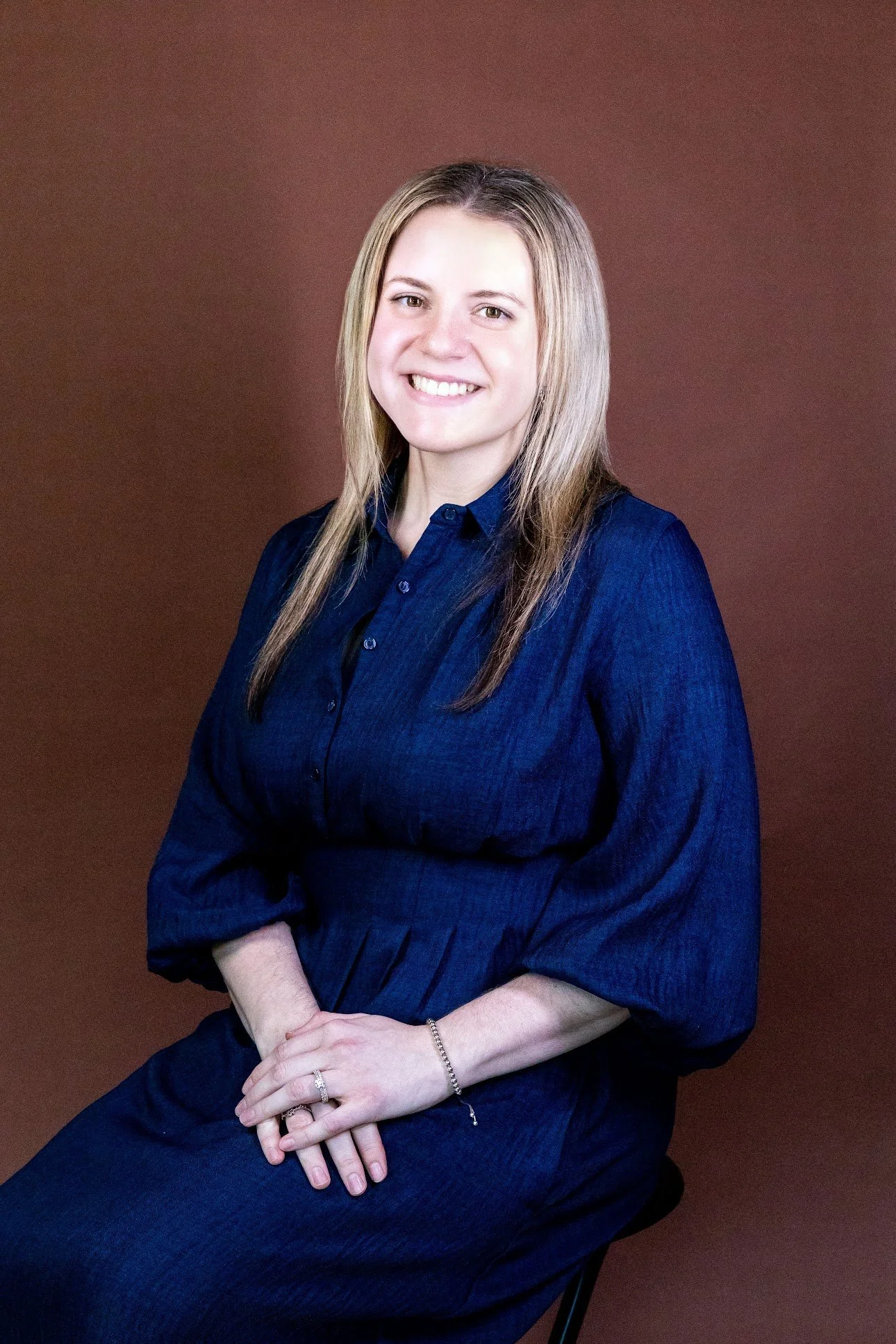 A woman with shoulder-length blonde hair, wearing a dark blue button-up dress, sitting against a plain brown background, smiling with her hands clasped.