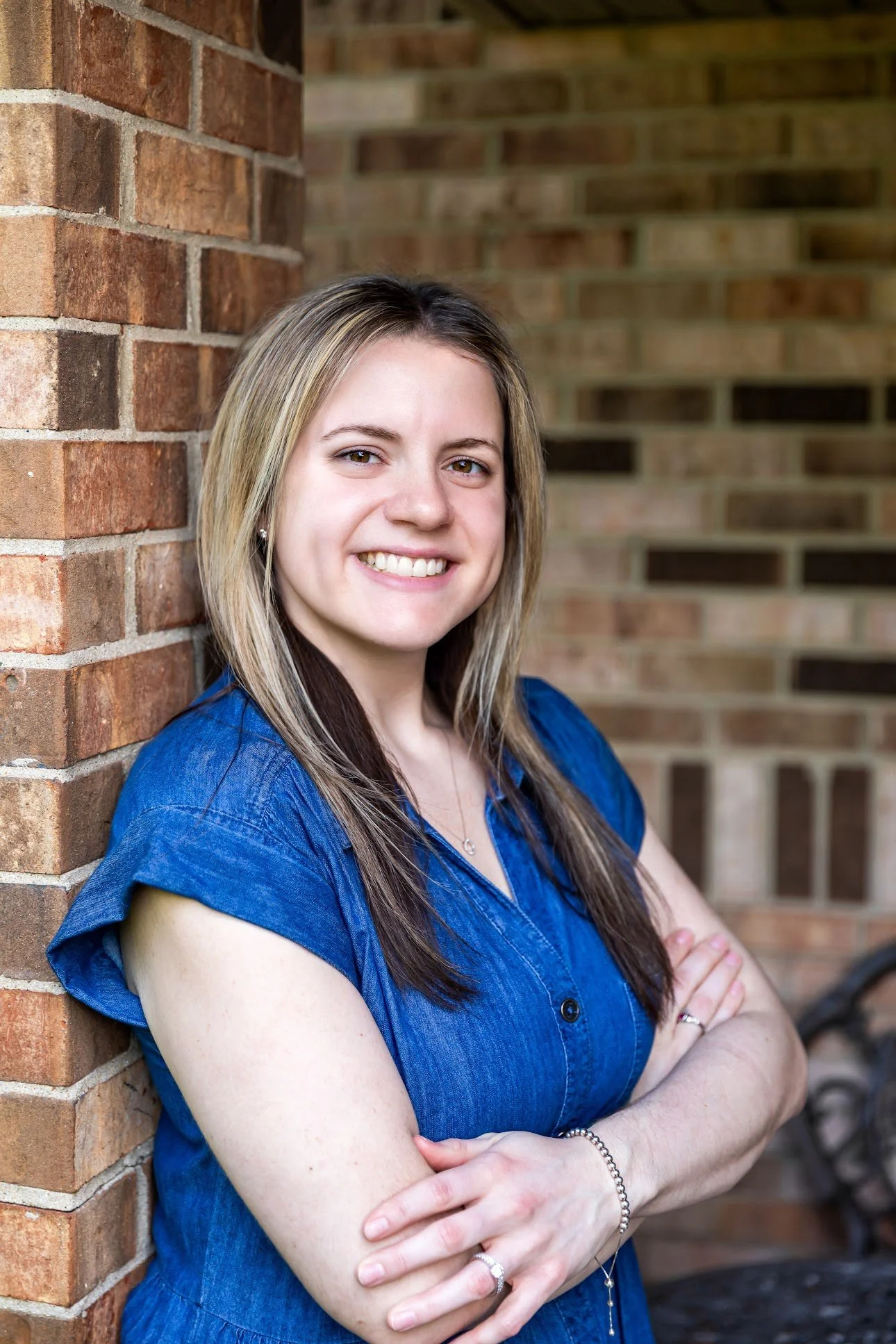 A woman with long, light brown hair and a blue shirt, smiling and standing with arms crossed in front of a brick wall.
