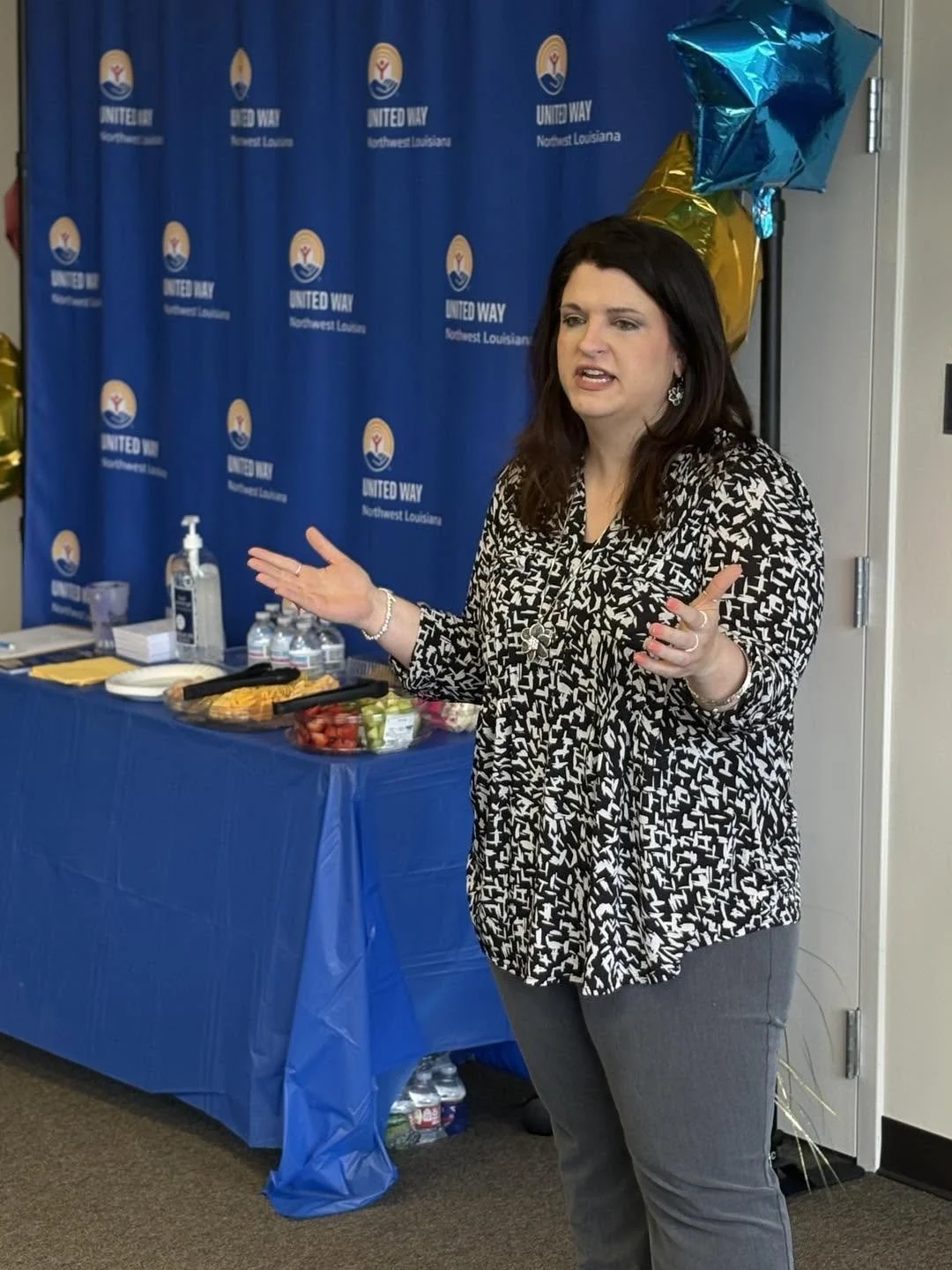 A woman speaking in front of a blue United Way backdrop with balloons and a table of snacks, including fruit, hand sanitizer, and water bottles.