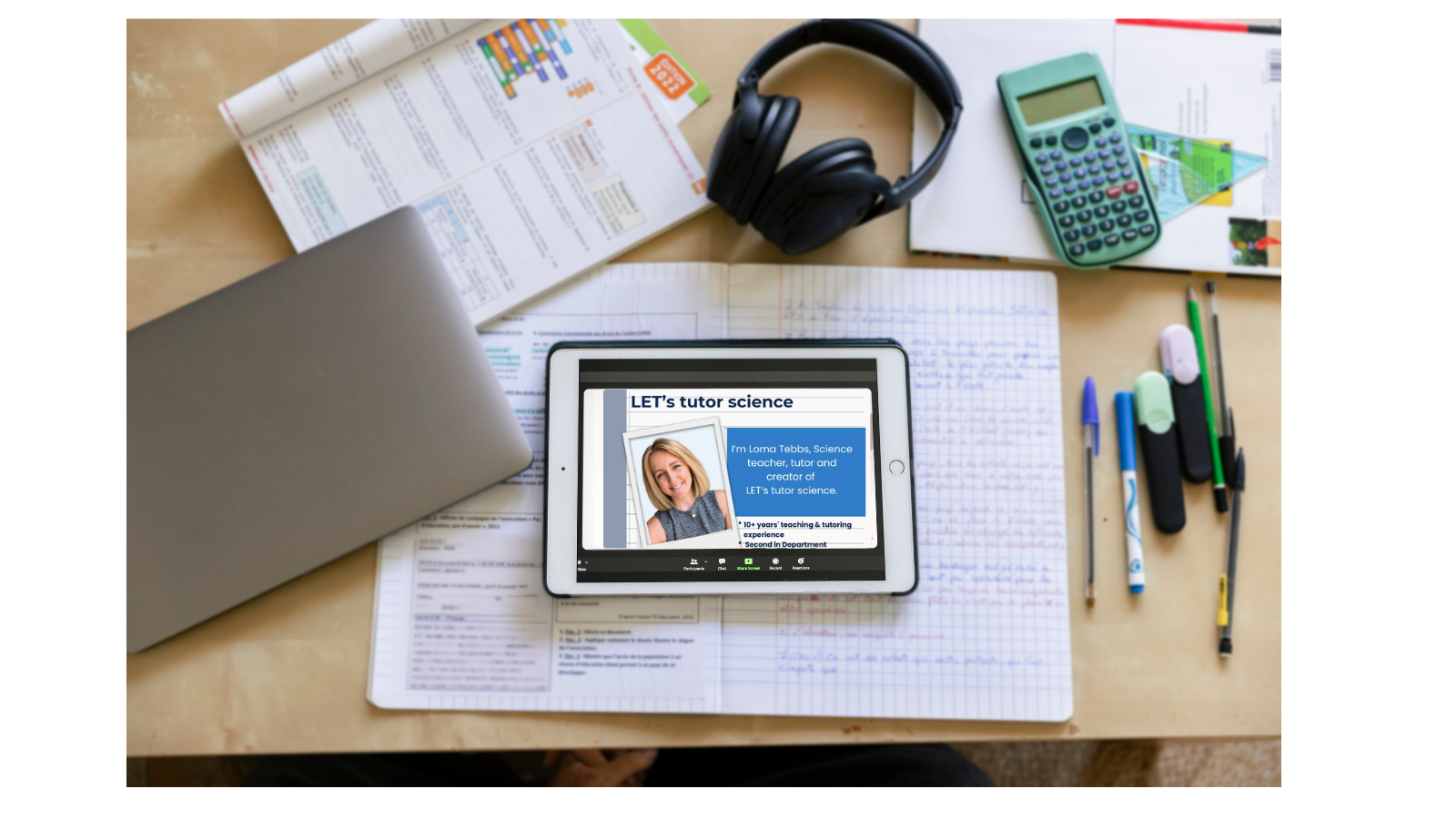 A study desk with a closed silver laptop, an open graph paper notebook with handwritten notes, a tablet displaying a tutor profile, a pair of headphones, a calculator, a protractor, pens, and highlighters on top of documents and papers.