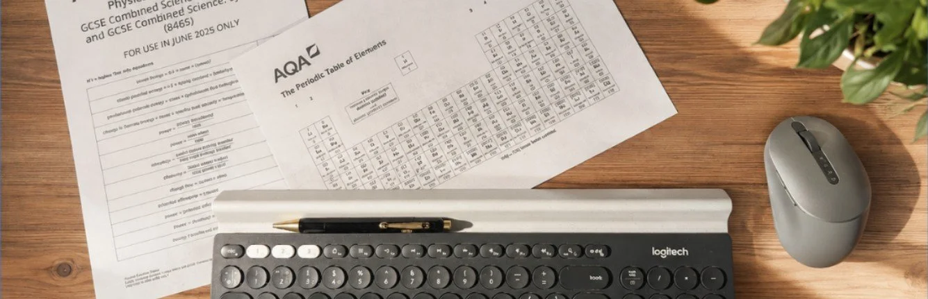 Desk with a paper on the periodic table, a computer keyboard, a wireless mouse, a pen, and a plant.