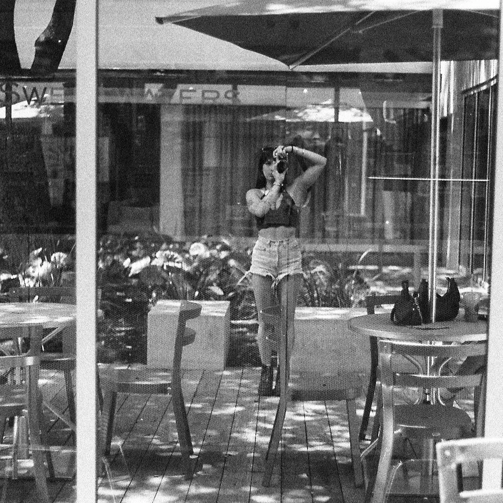 A woman taking a selfie in a cafe window. She is wearing a black top and denim shorts. The reflection shows trees, outdoor seating, and motorcycles outside.