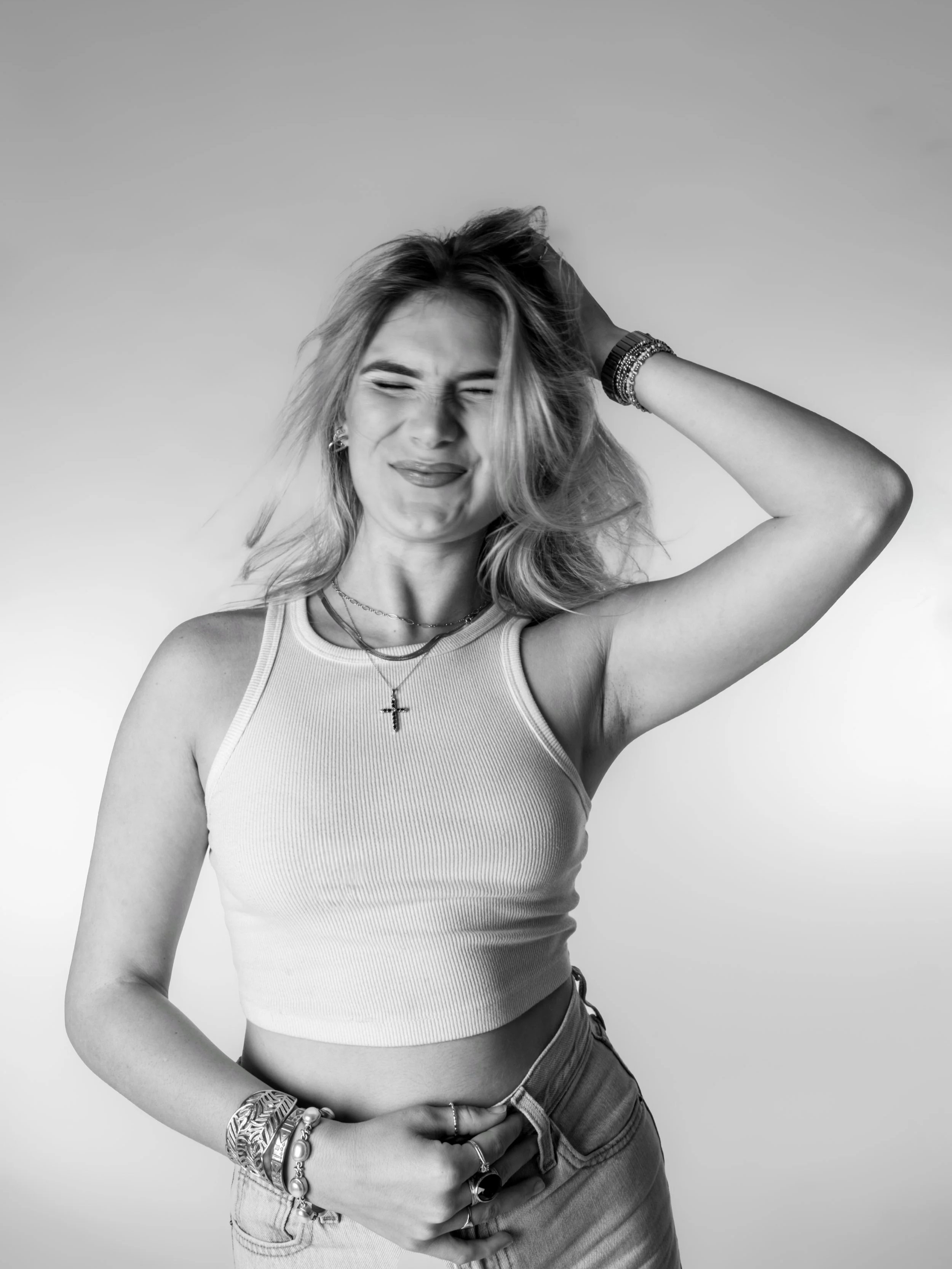 Black and white photo of a young woman with wavy hair, wearing a sleeveless top, multiple bracelets, a cross necklace, and rings, with her eyes closed and a pained or distressed expression, holding her head with one hand.