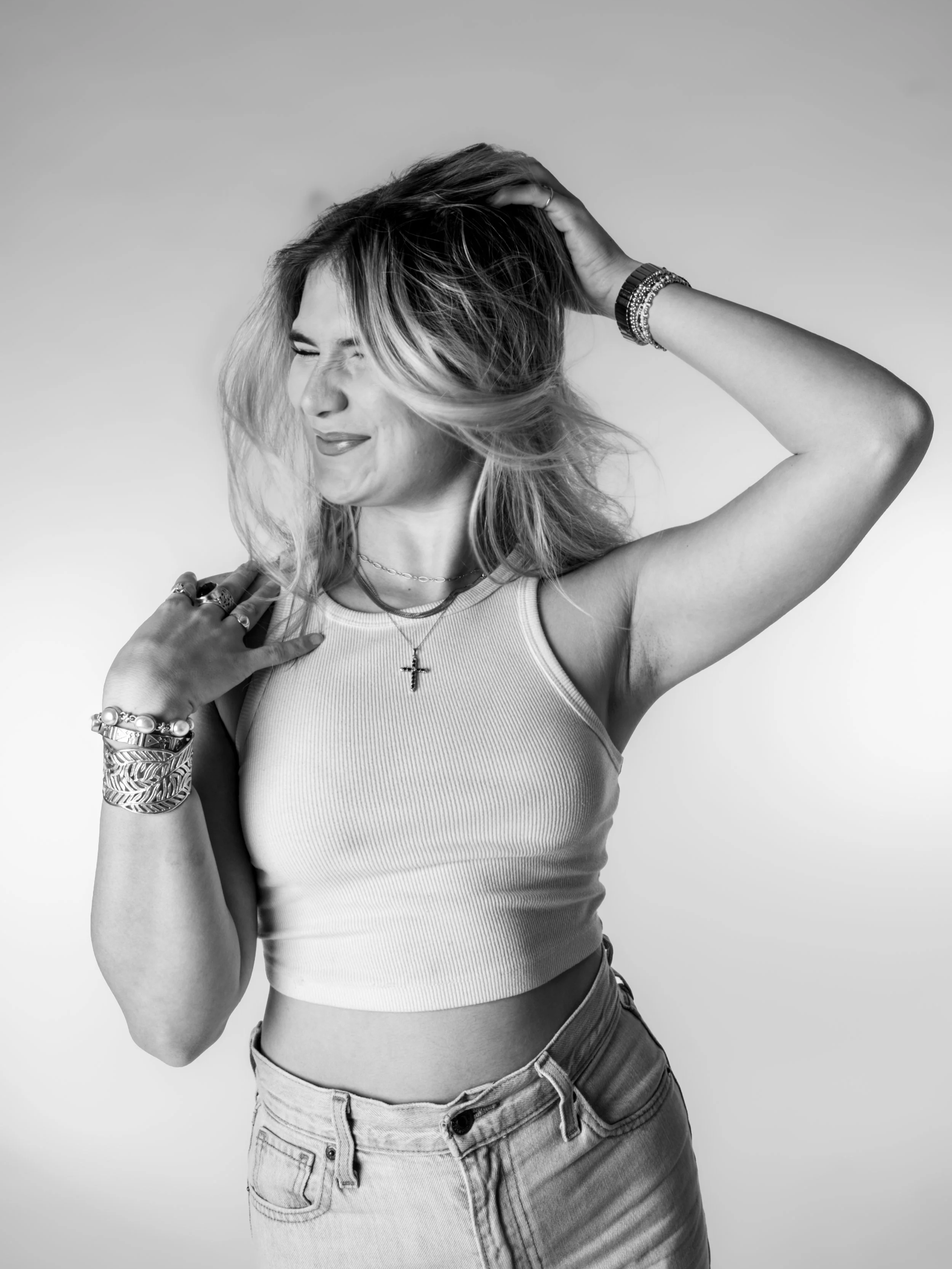 Black and white photo of a woman smiling, with her eyes closed, holding her hair, wearing a sleeveless top, multiple bracelets, rings, necklace with a cross pendant, and high-waisted jeans.