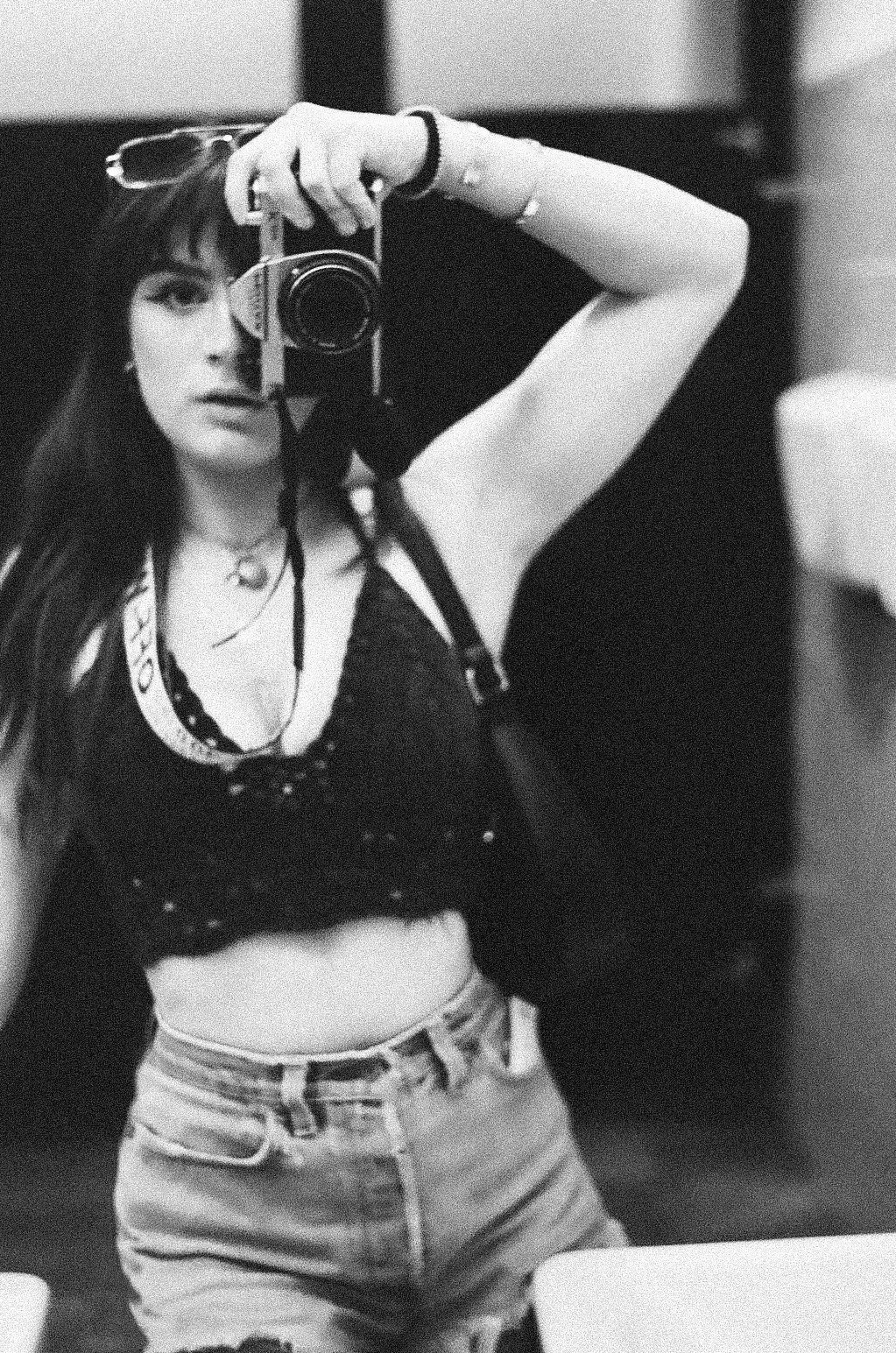 Black and white photo of a woman taking a mirror selfie. She has long hair with bangs and is wearing glasses, a crop top, and high-waisted shorts.