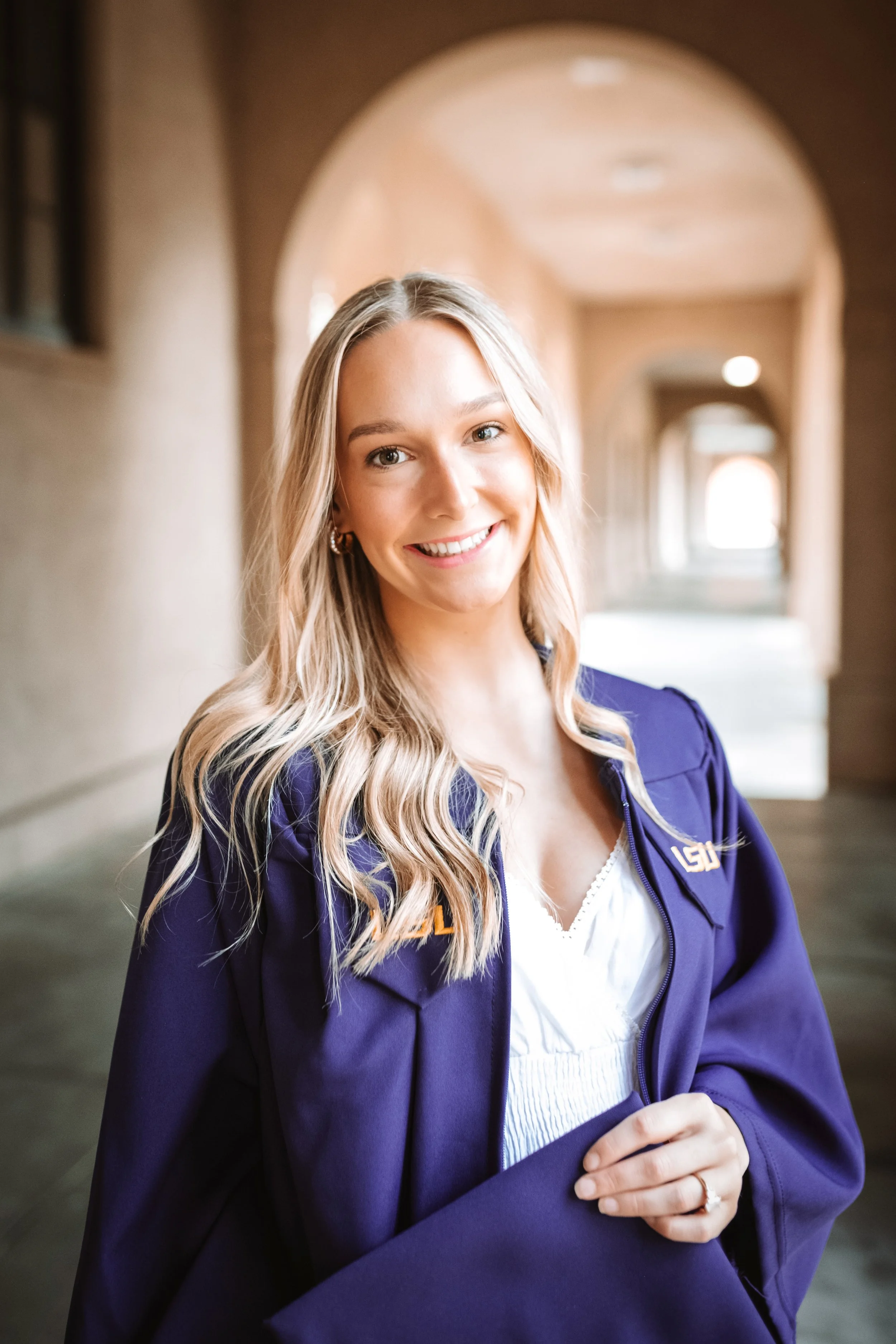 Young woman with long blond hair smiling in a corridor with arches, wearing a purple jacket with a University of Louisiana at Lafayette logo.
