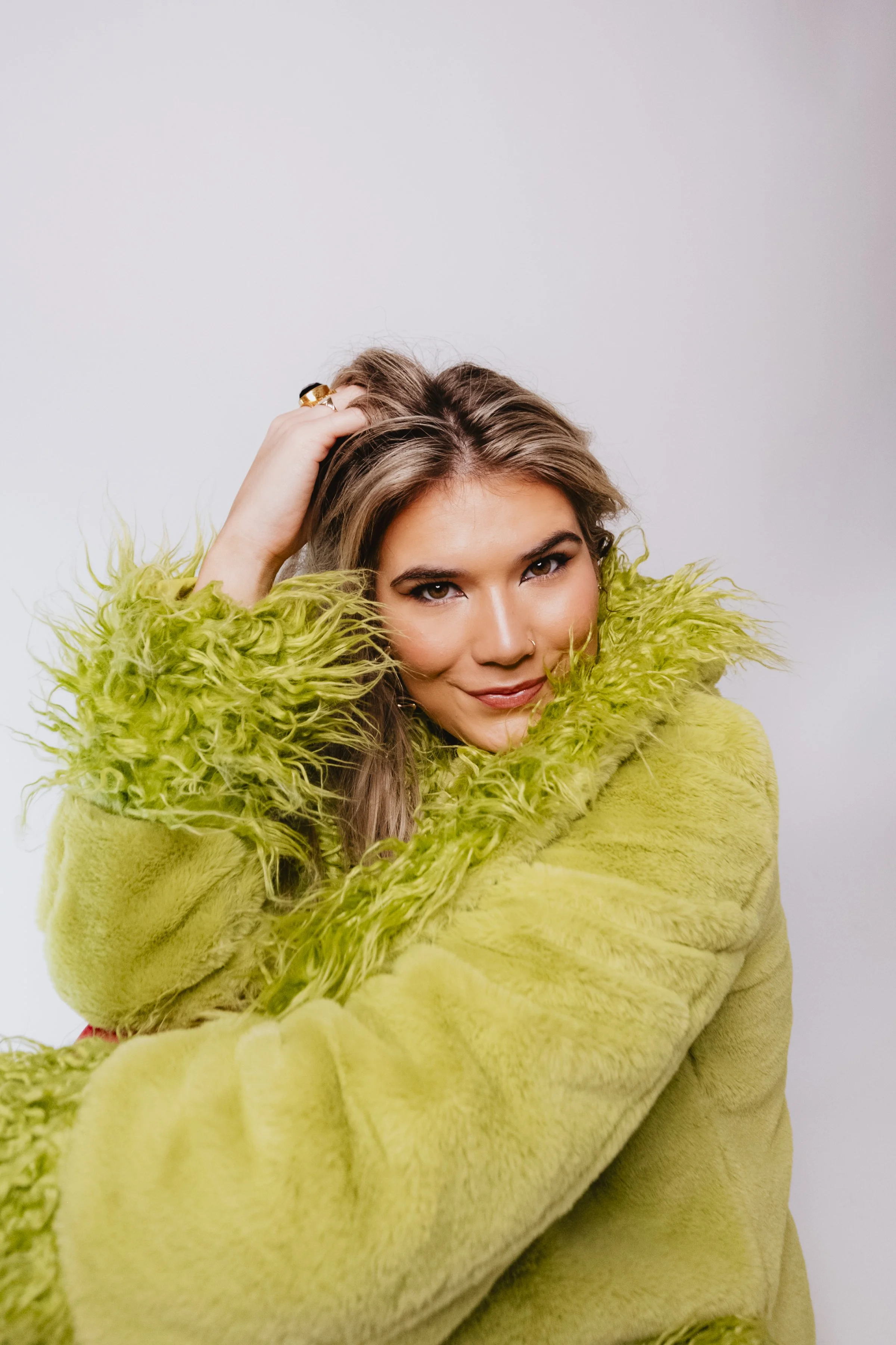 A woman with brown hair, brown eyes, and light skin, wearing a bright green fuzzy coat with a large collar, smiling and looking at the camera, in front of a plain white background.