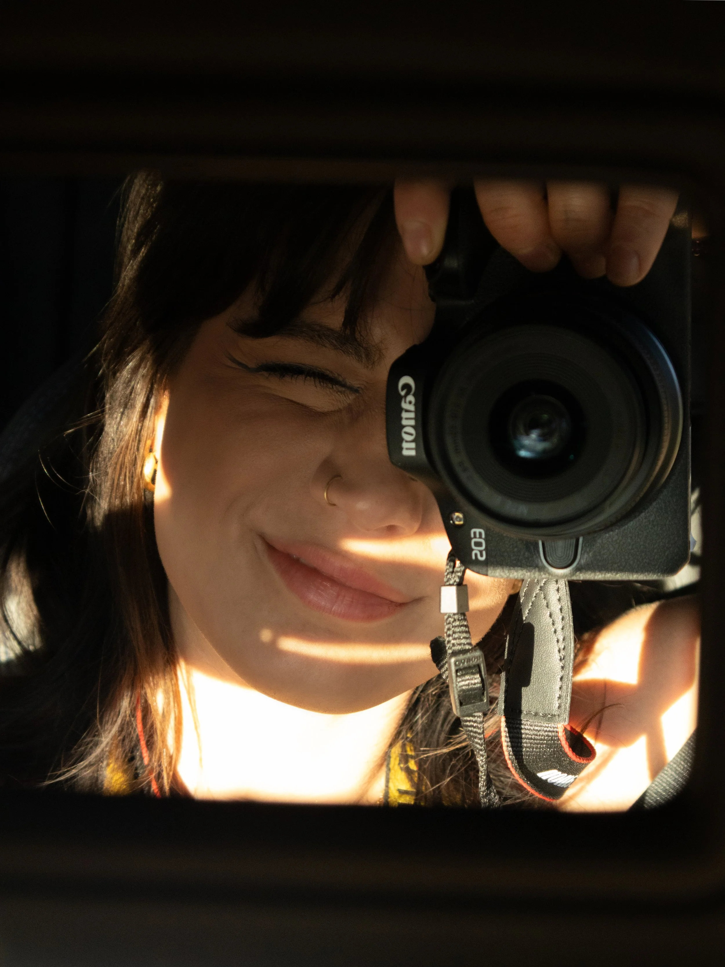 A person taking a self-portrait with a Canon EOS camera, showing part of their face with closed eyes and long dark hair, in a partially illuminated environment.