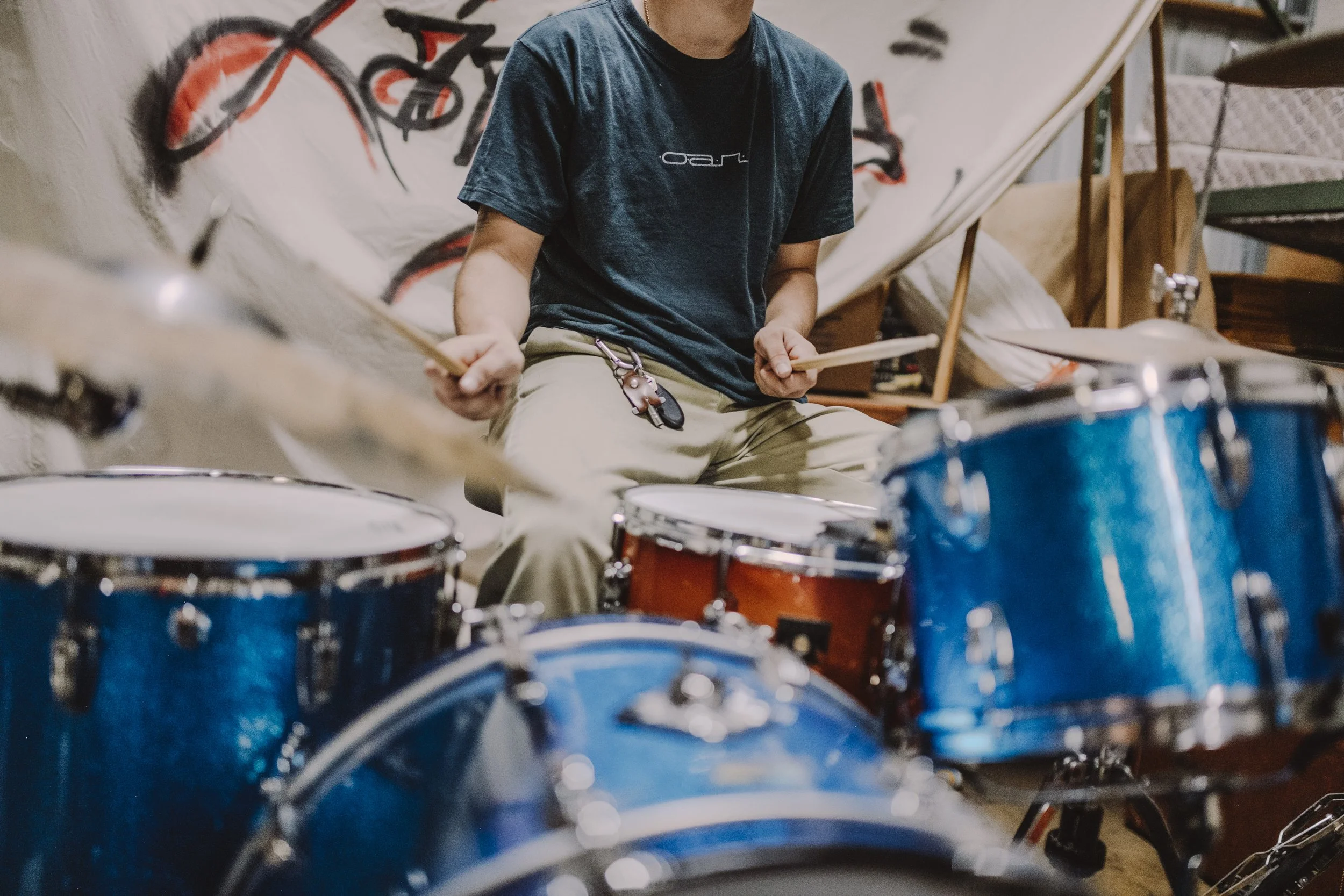 A person wearing a dark blue t-shirt and beige pants sitting on a sofa, holding drumsticks, surrounded by drums and other musical equipment in a room with graffiti on the wall.