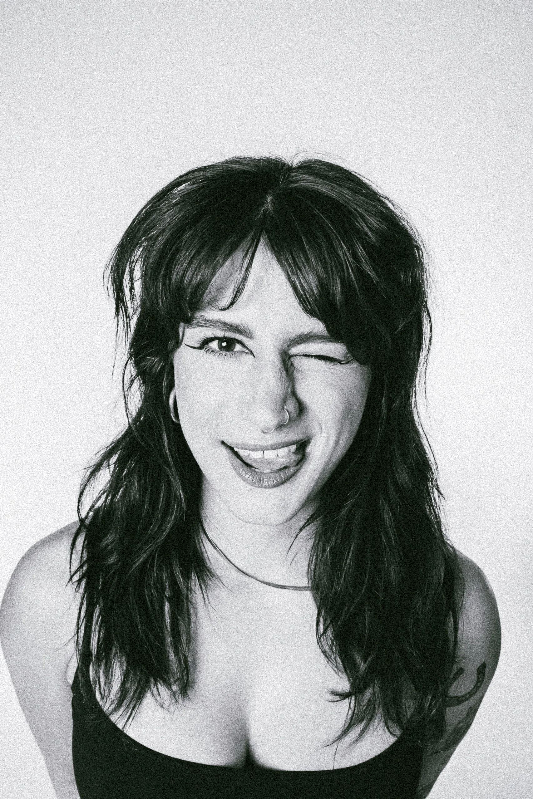 Black and white photo of a woman with dark, wavy hair, winking and smiling with tongue slightly out, wearing a dark top, against a plain light background.