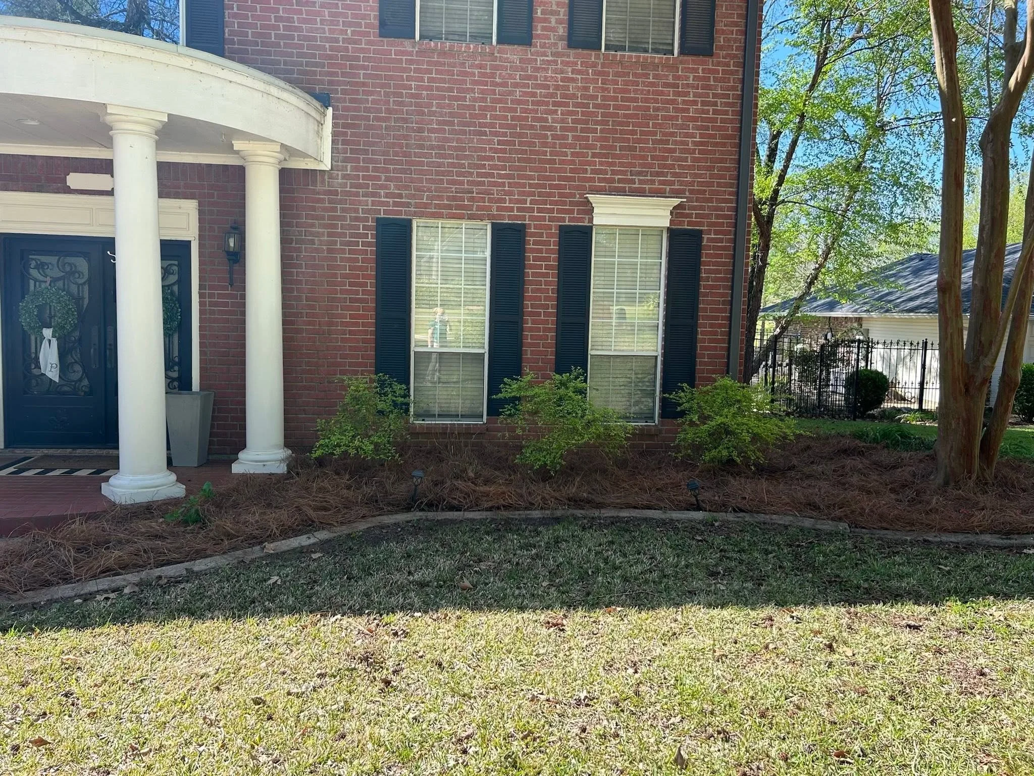 A residential house with a brick exterior, black window shutters, and white columns at the front entrance. There is a small front yard with bushes, trees, and grass, and a clear, sunny sky.