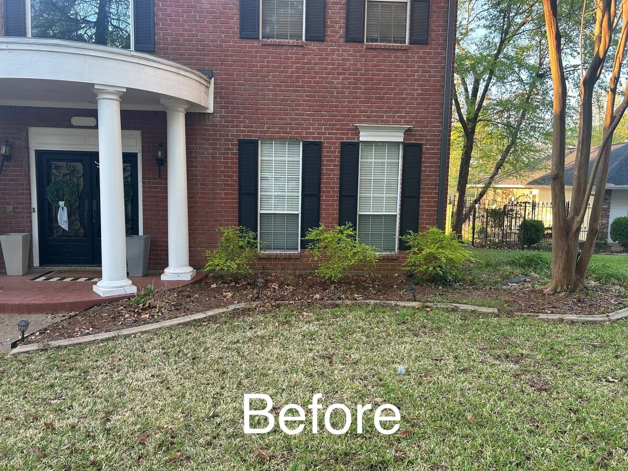 Front view of a brick house with black shutters, white columns, and a lawn with small bushes and a tree.
