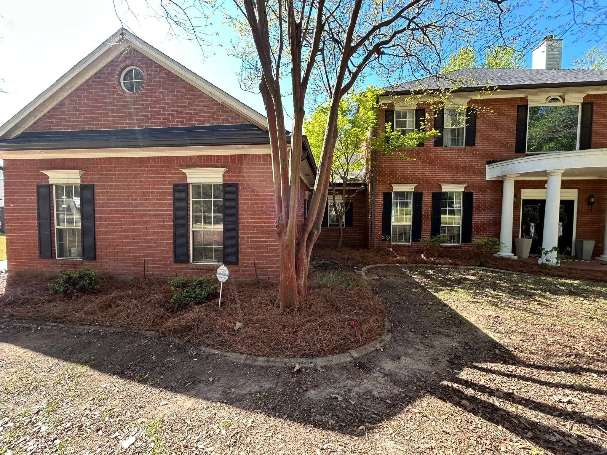 Front view of a brick house with a gable roof and a large window, surrounded by trees and a garden area with mulch and plants.