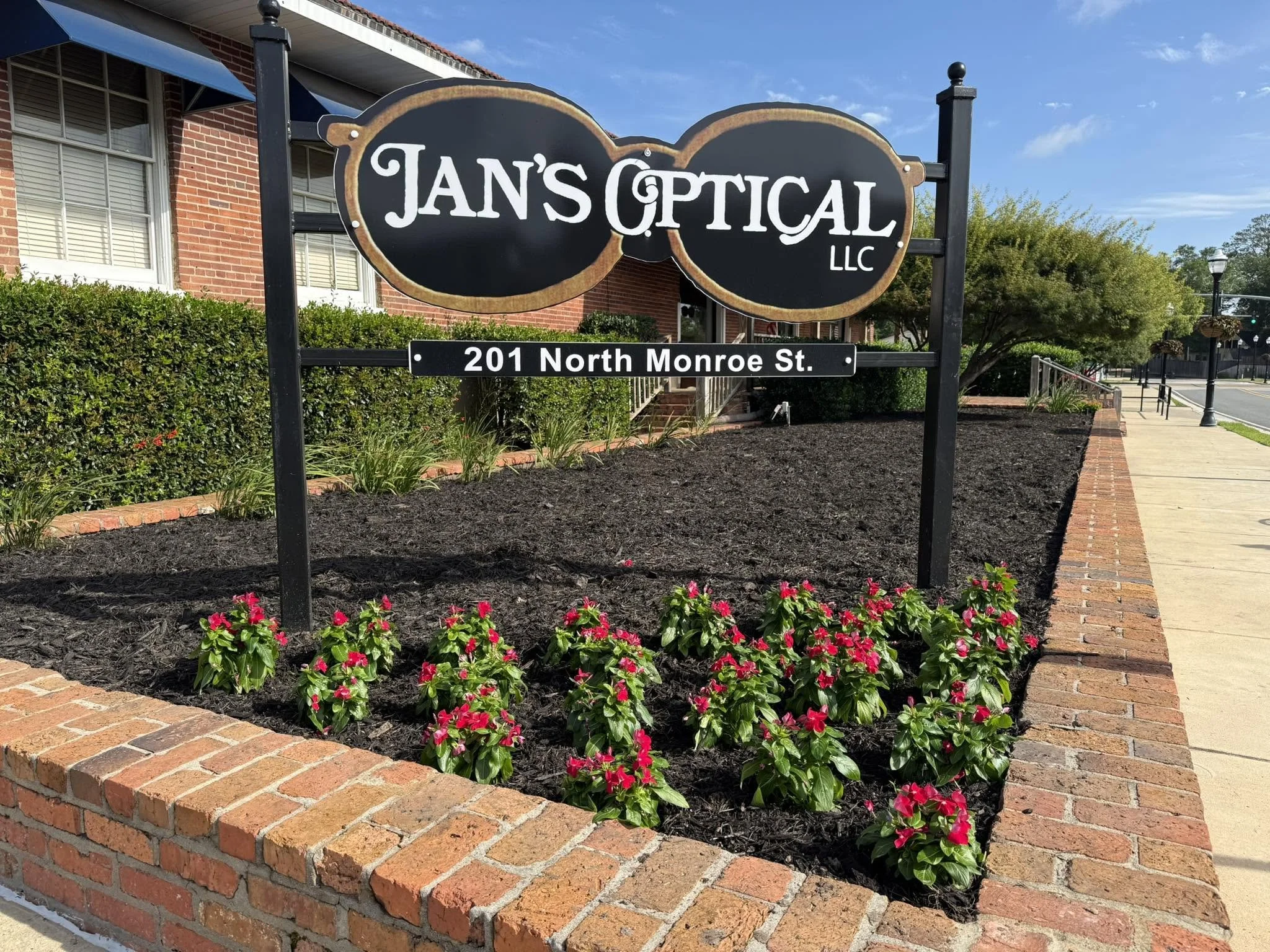 Sign for Jan's Optical LLC located at 201 North Monroe Street, with a brick building, a sidewalk, and trees in the background.