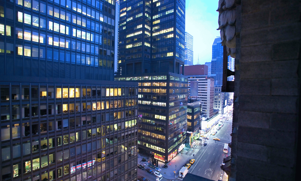 View of a city street at dusk with tall buildings, some with illuminated windows, and a few cars on the road.