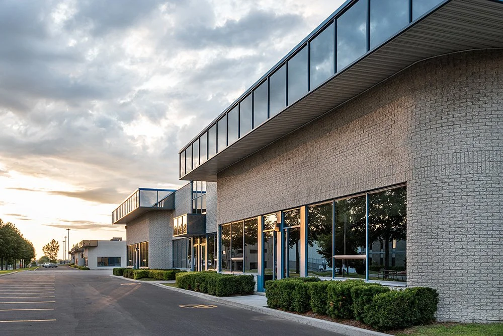 Modern commercial building with large glass windows, beige brick exterior, and a parking lot in front at sunset.