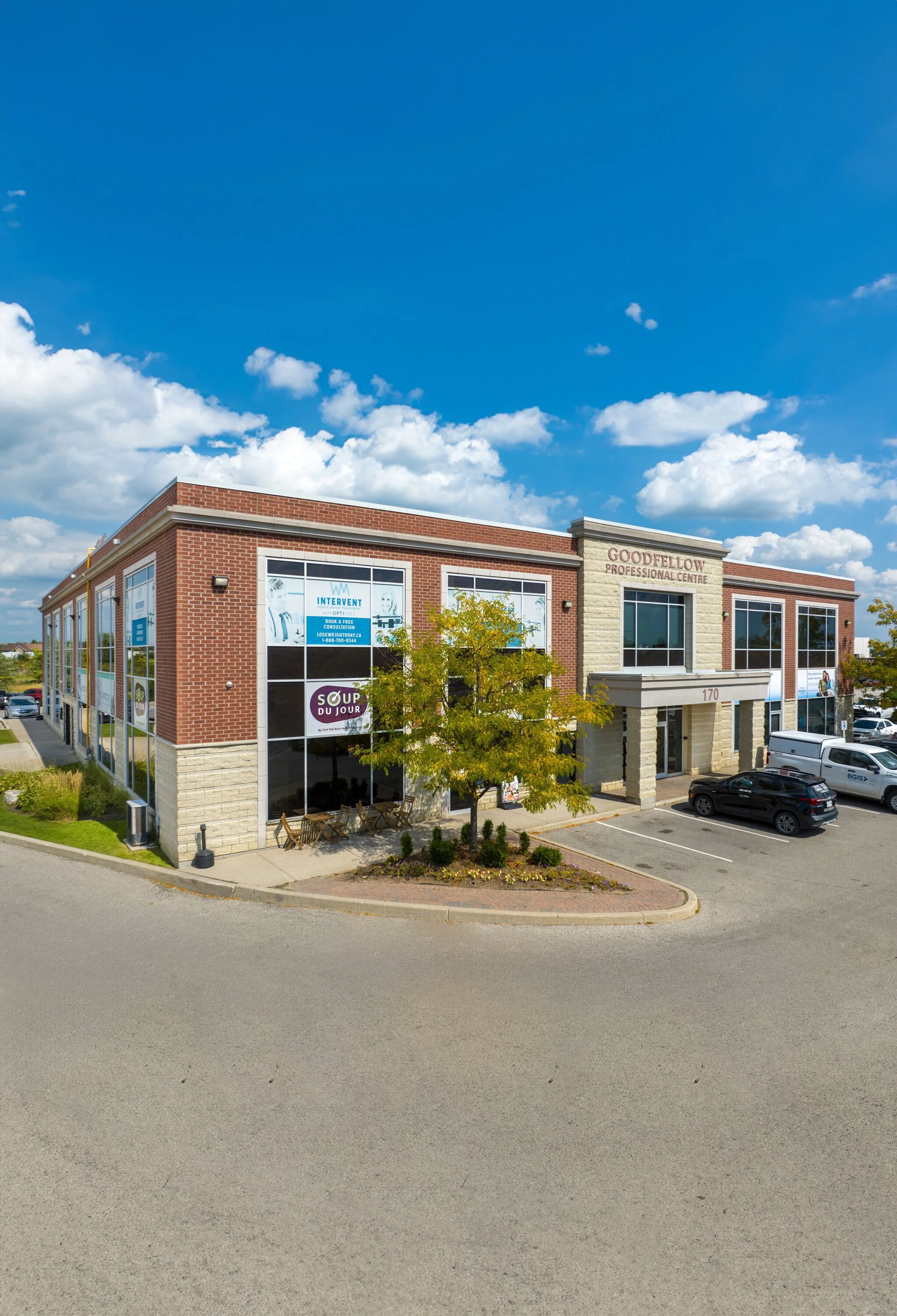 A modern two-story commercial building with brick and stone exterior, large windows, and a sign reading 'Goodfellow Professional Centre.' There are parking spaces in front, a tree with green leaves, and a blue sky with scattered clouds overhead.