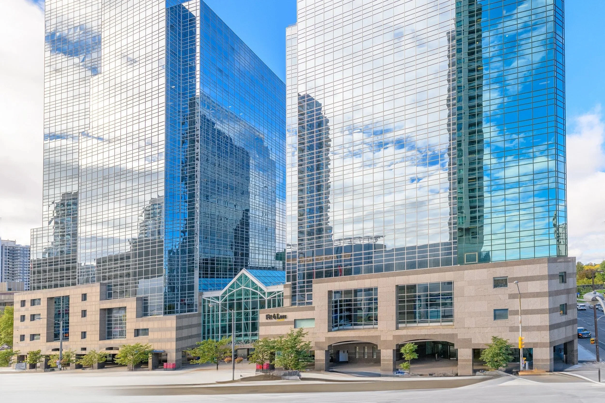 Modern office building with a glass facade reflecting the sky and nearby structures, with a lower beige stone section and a small parking lot in front.