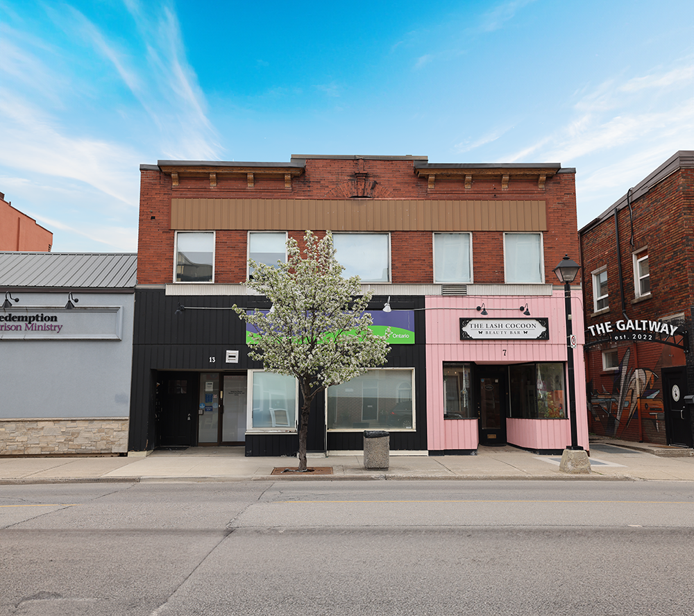 Street view of a three-story building with a tree in front, featuring a pink storefront for The Lash Cocoon beauty bar and a black storefront for a business, under a blue sky with wispy clouds.