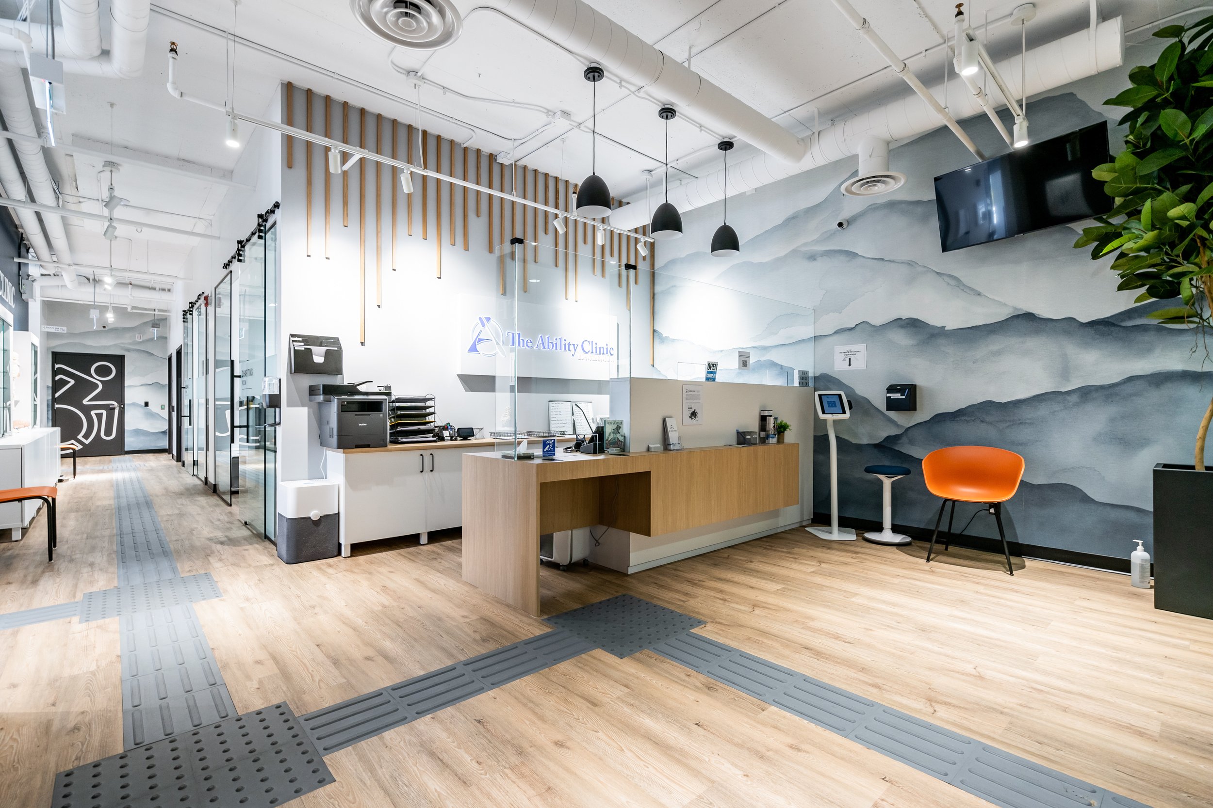 Interior of a modern medical clinic reception area with a wooden counter, a digital check-in kiosk, orange chairs, a large wall mural of mountains, and visible ceiling piping.