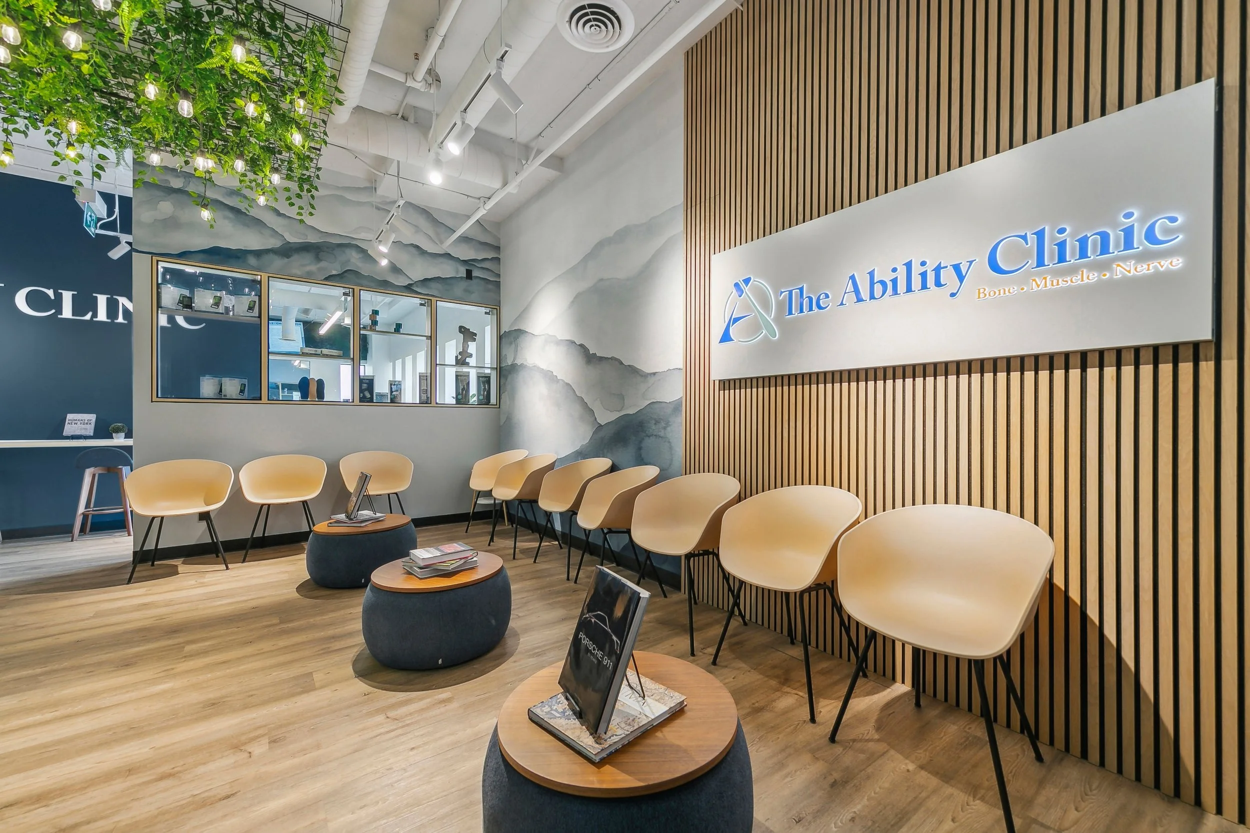 Waiting area of The Ability Clinic with beige chairs, round tables displaying magazines and brochures, a wall with a sign reading 'The Ability Clinic' and ‘Bone, Muscle, Nerve,’ a background mural of mountains, a window with visible office interior, and decorative hanging greenery.