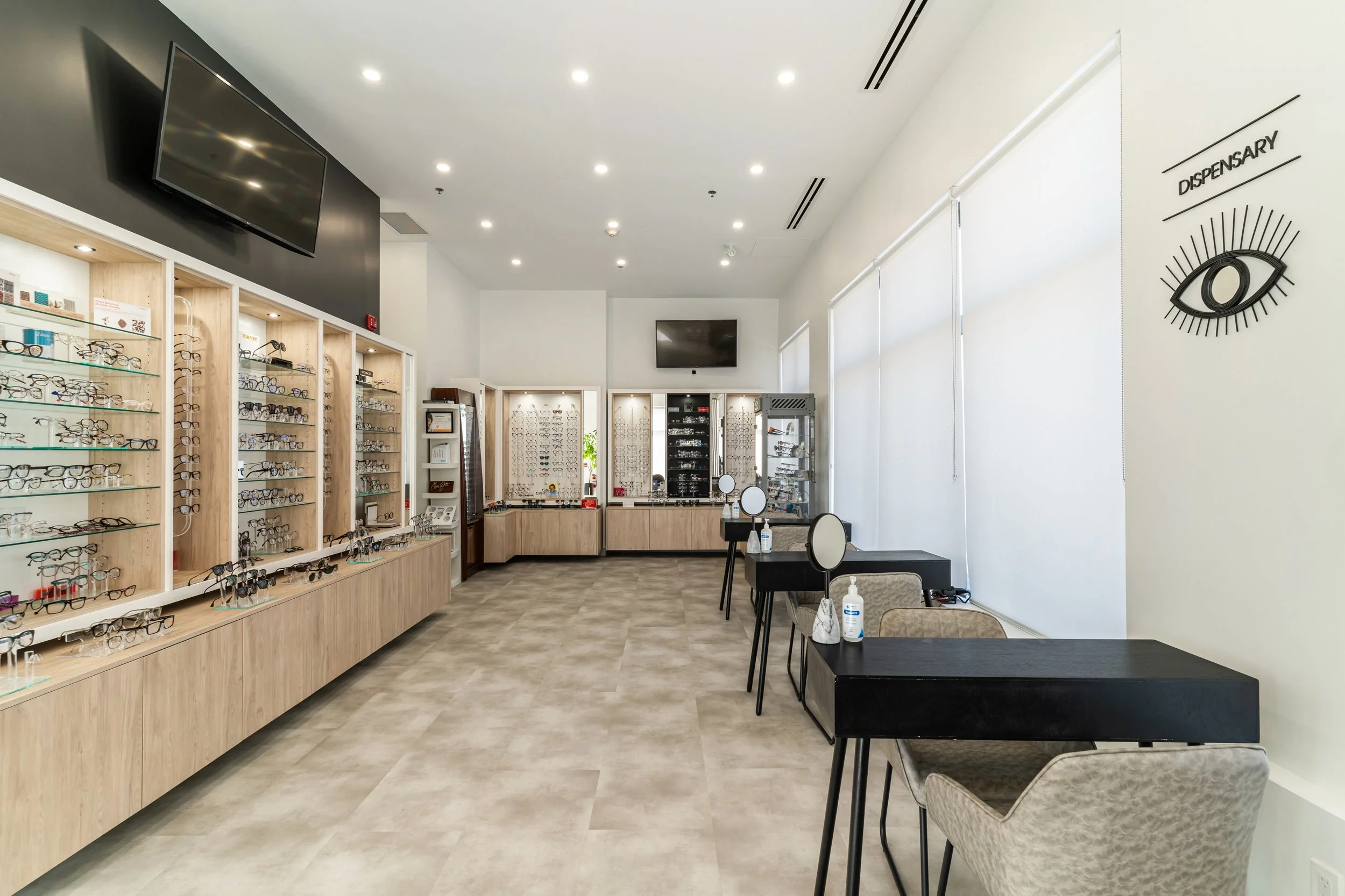 Interior of an optometry dispensary with display shelves of eyeglasses, make-up stations, and a sign with an eye logo that says 'Dispensary'.