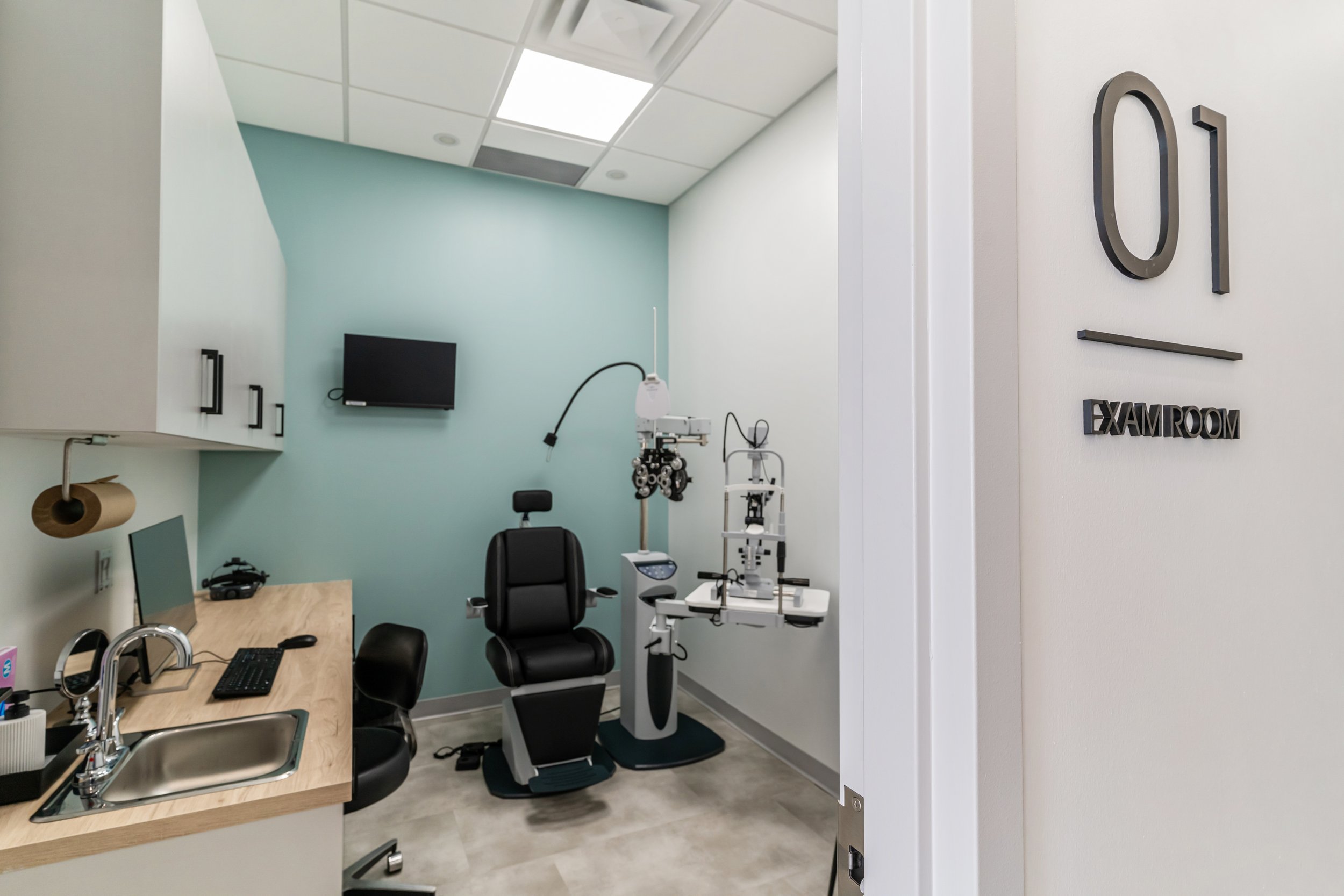 Exam room with medical examination chair, eye examination equipment, small desk, and wall-mounted TV in a modern medical facility.