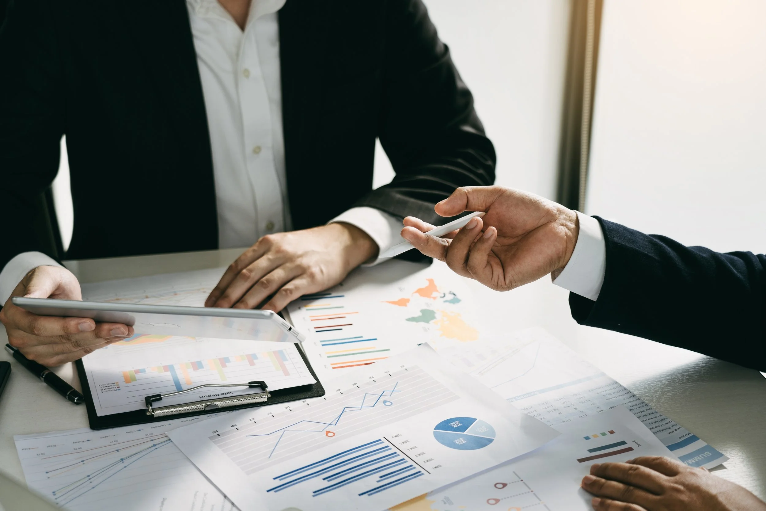 Two people are engaged in a business discussion at a desk covered with financial charts and graphs. One person is showing data on a tablet, while the other is pointing at the documents with charts and graphs.