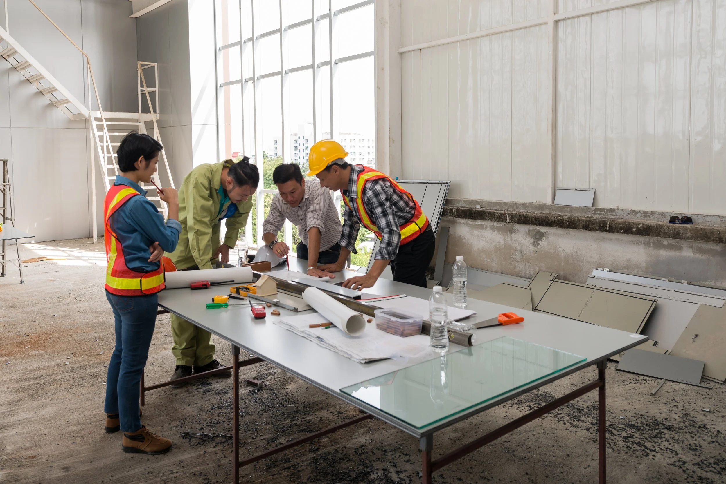 Group of five construction professionals, two men and three women, gathered around a table with blueprints and tools at a construction site. They are discussing project plans inside a building under construction.